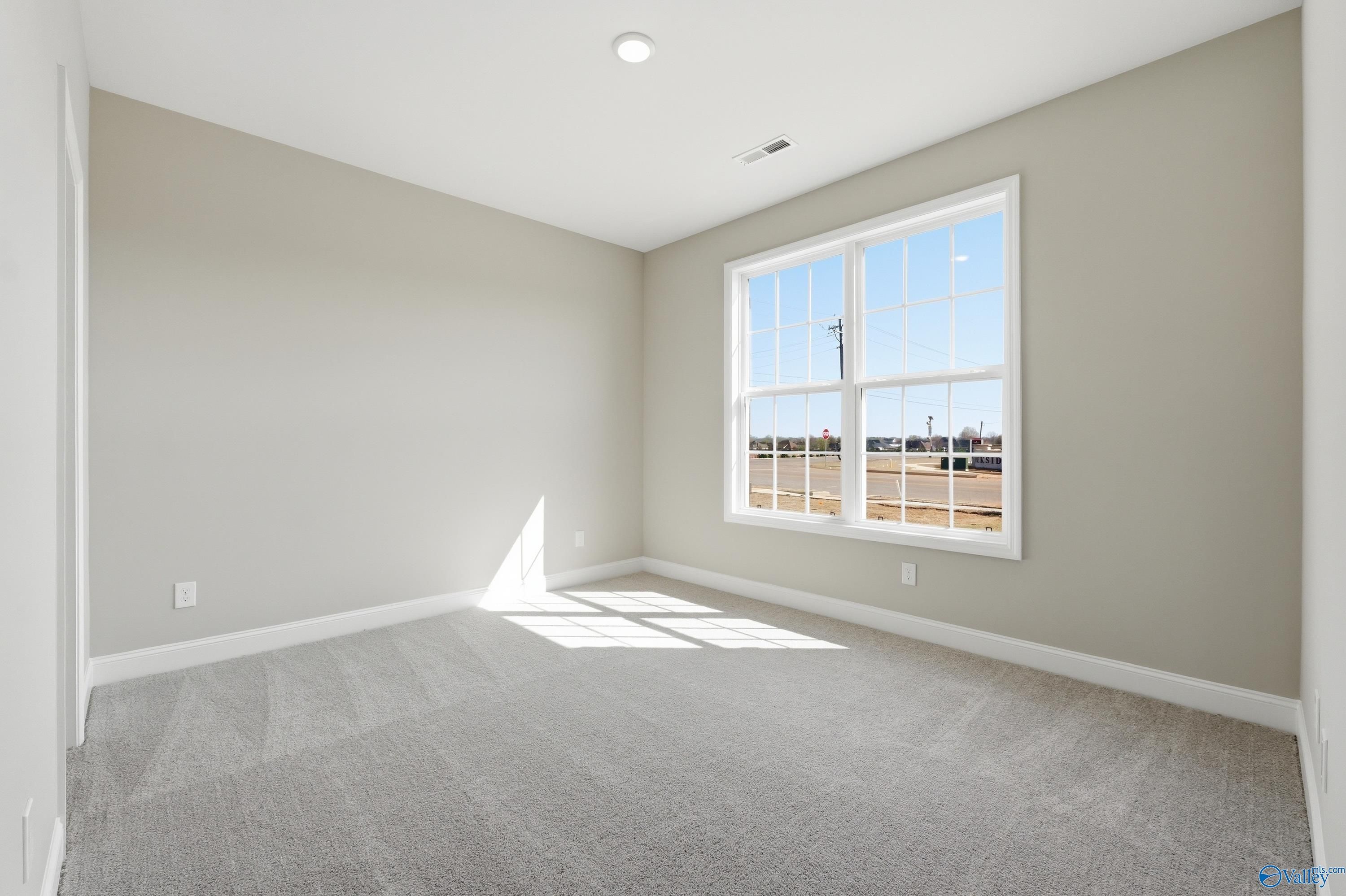 Bright empty bedroom with beige walls, gray carpet, and large sunny window in Davidson Homes The Rockford, Harvest, Alabama