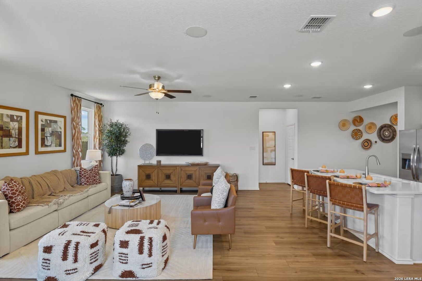 Open-concept living room with beige sectional sofa, TV console, and kitchen island with bar stools in Davidson Homes The Daphne K, Bricewood, San Antonio