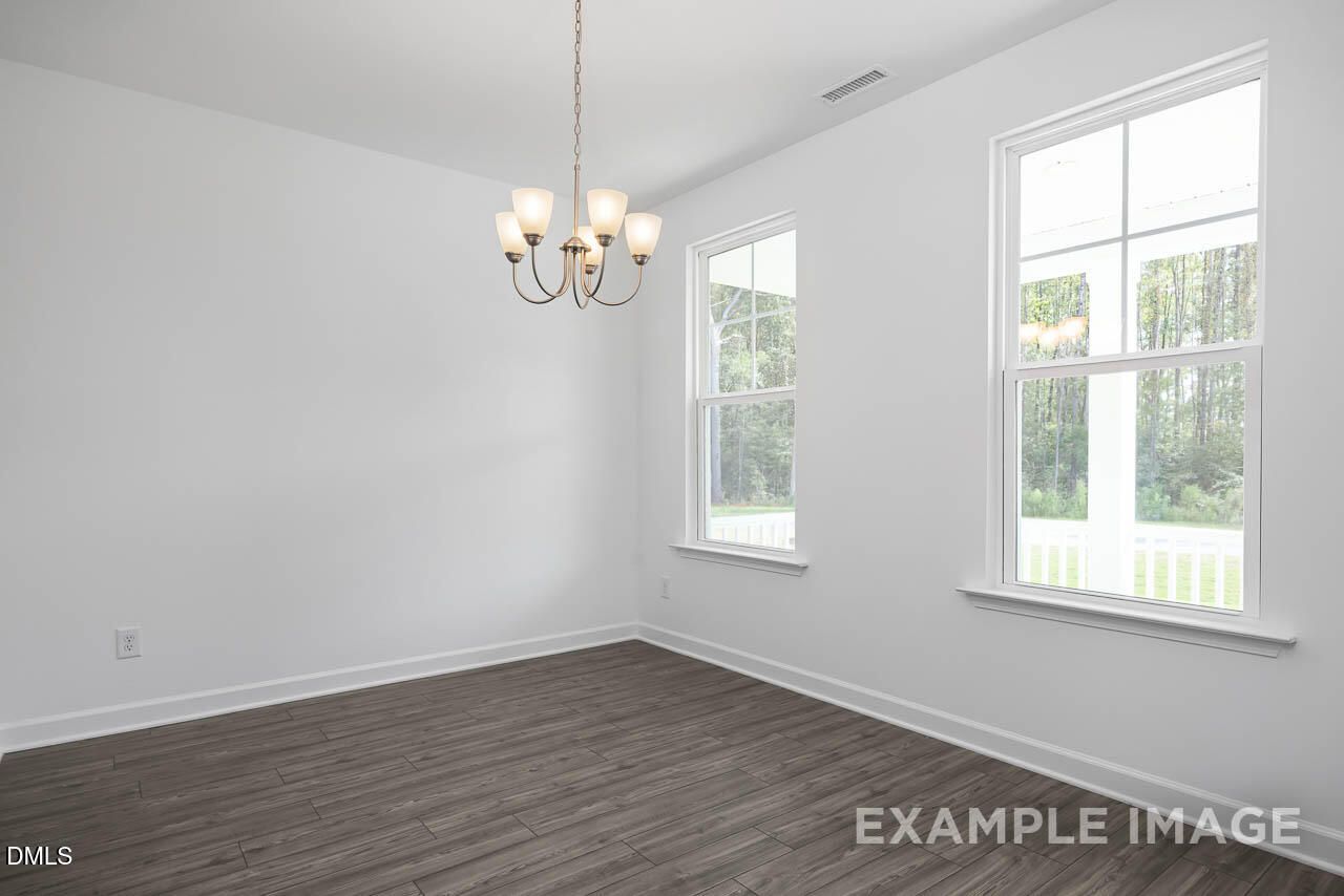 Bright empty living room with hardwood floors, chandelier, and large windows overlooking woods in The Willow D, Zebulon, NC