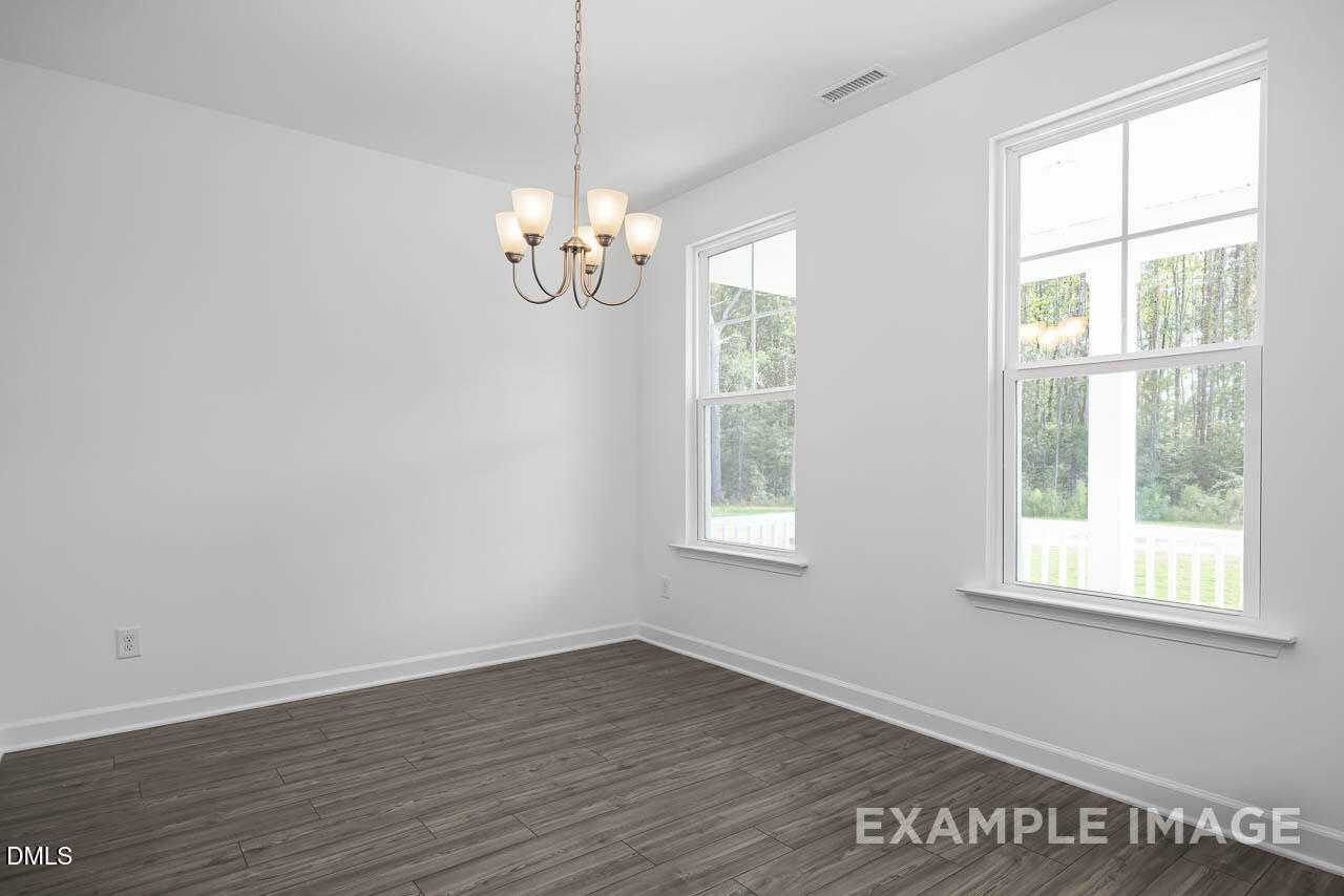 Bright empty living room with hardwood floors, chandelier, and large windows overlooking woods in The Willow D, Zebulon, NC