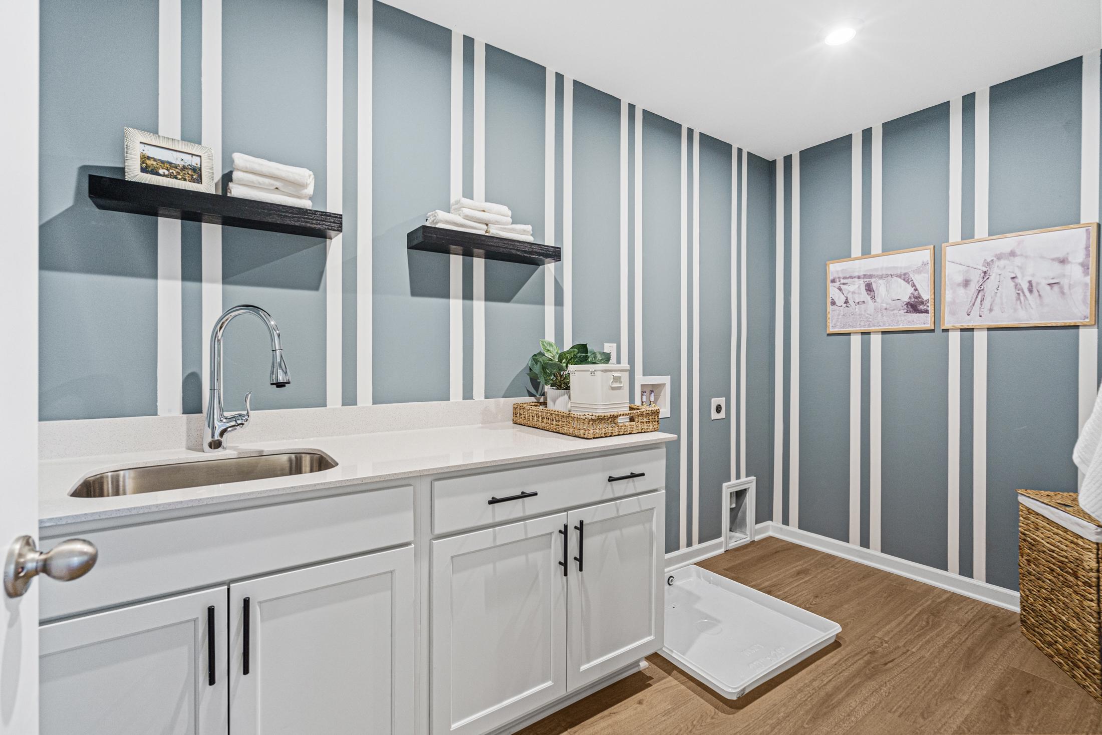 Modern laundry room in The Hickory B featuring blue striped walls, white shaker cabinets, deep utility sink, and floating shelves