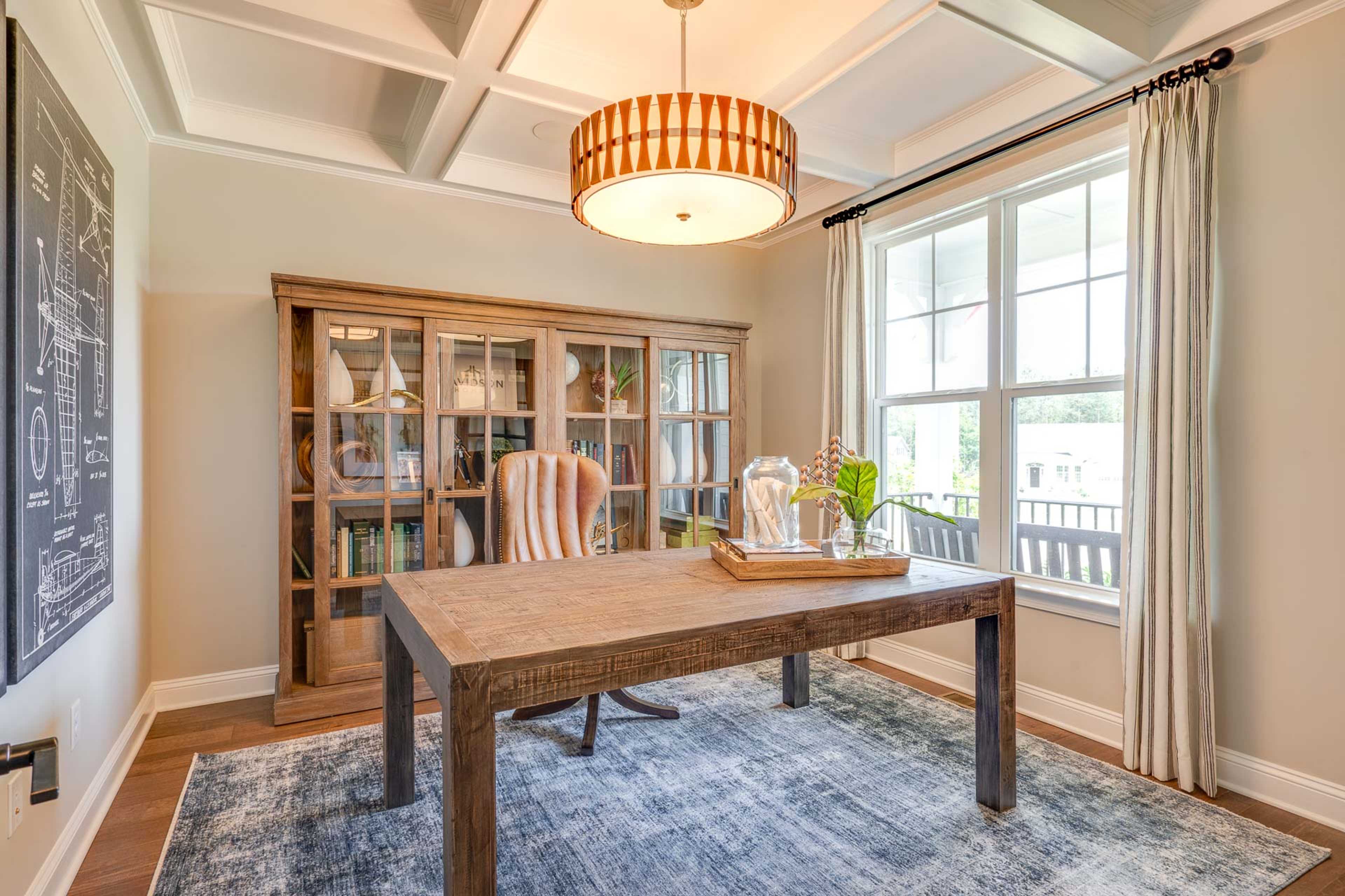 Cozy home office in Hasentree Wake Forest NC with wooden desk, glass-door bookshelves, chandelier, and large windows