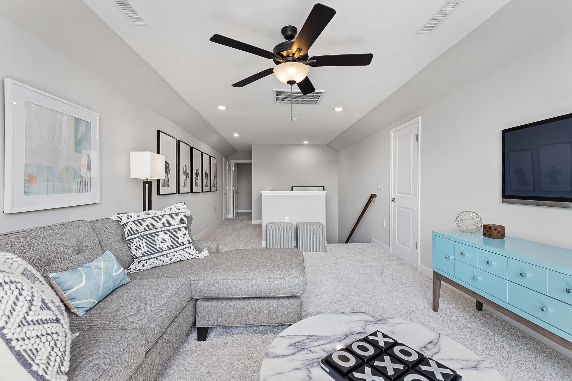 Spacious bonus loft in Highland Forest home by Davidson Homes, Fuquay-Varina NC with gray L-shaped sofa, ceiling fan, wall TV