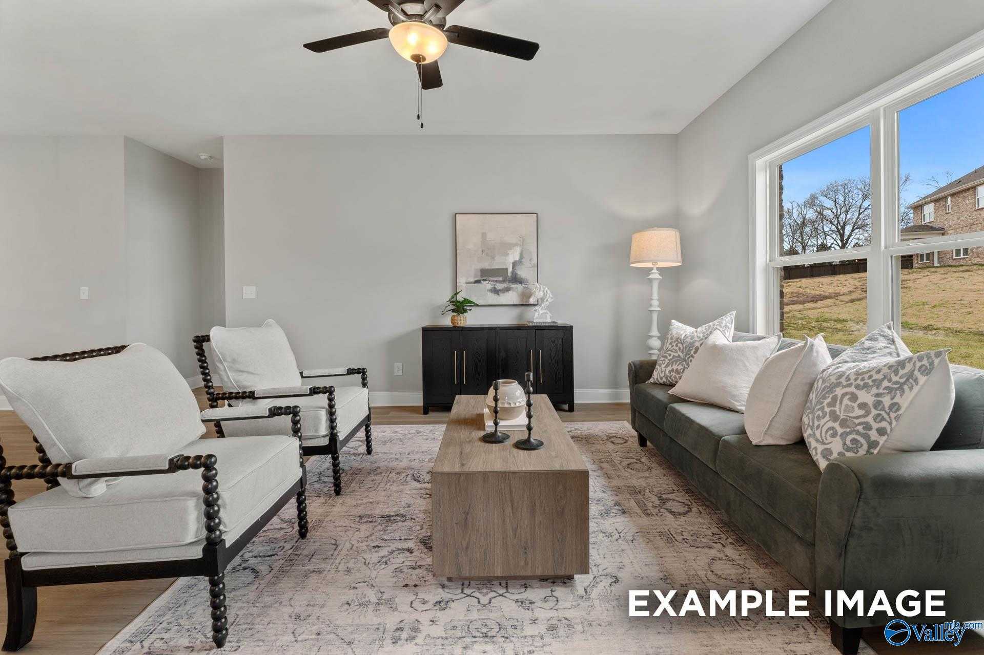 Inviting living room with gray sofa, white armchairs, ceiling fan, and large windows overlooking landscape in Davidson Homes The Daphne C, Owens Cross Roads, Alabama
