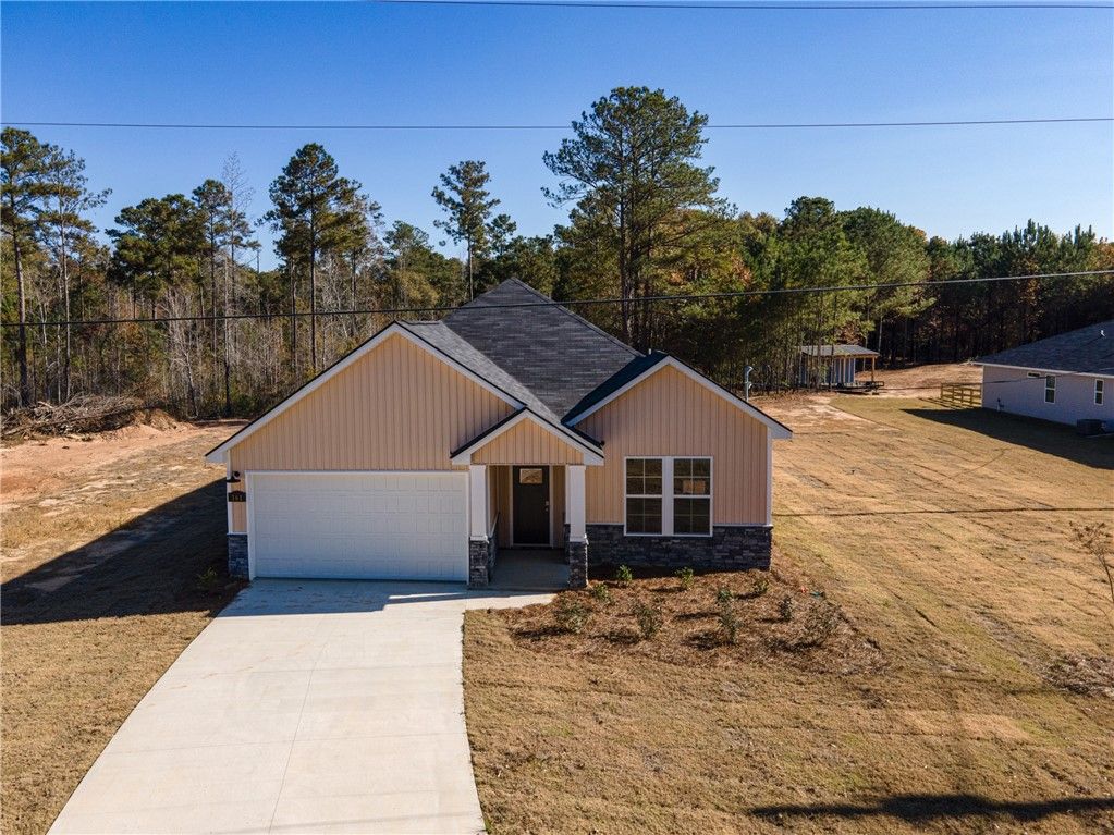 Single-story beige home with dark shingle roof, 1-car garage, and driveway amid pine trees in Silver Oak, Cusseta, Alabama
