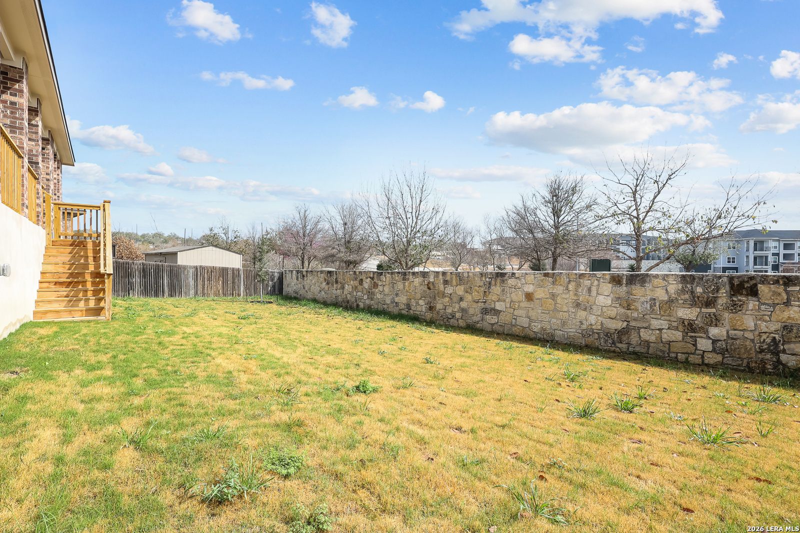 Spacious grassy backyard with stone wall, wooden deck stairs, and bare trees in Davidson Homes The Rockford G, Ladera, San Antonio