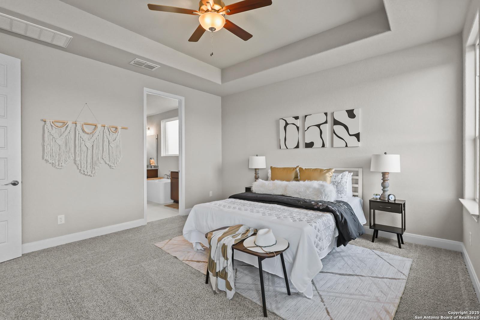 Serene master bedroom with tray ceiling, ceiling fan, king bed, abstract wall art, and en-suite bath in Davidson Homes The Jennings G, Castroville, Texas