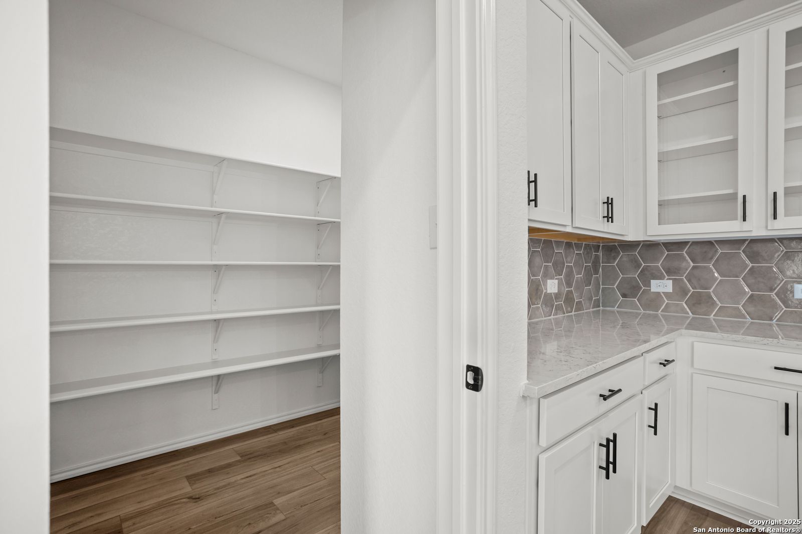 Modern white kitchen with quartz counters, gray hexagon backsplash, glass cabinets, and adjacent walk-in pantry in Davidson Homes The Daphne K, San Antonio