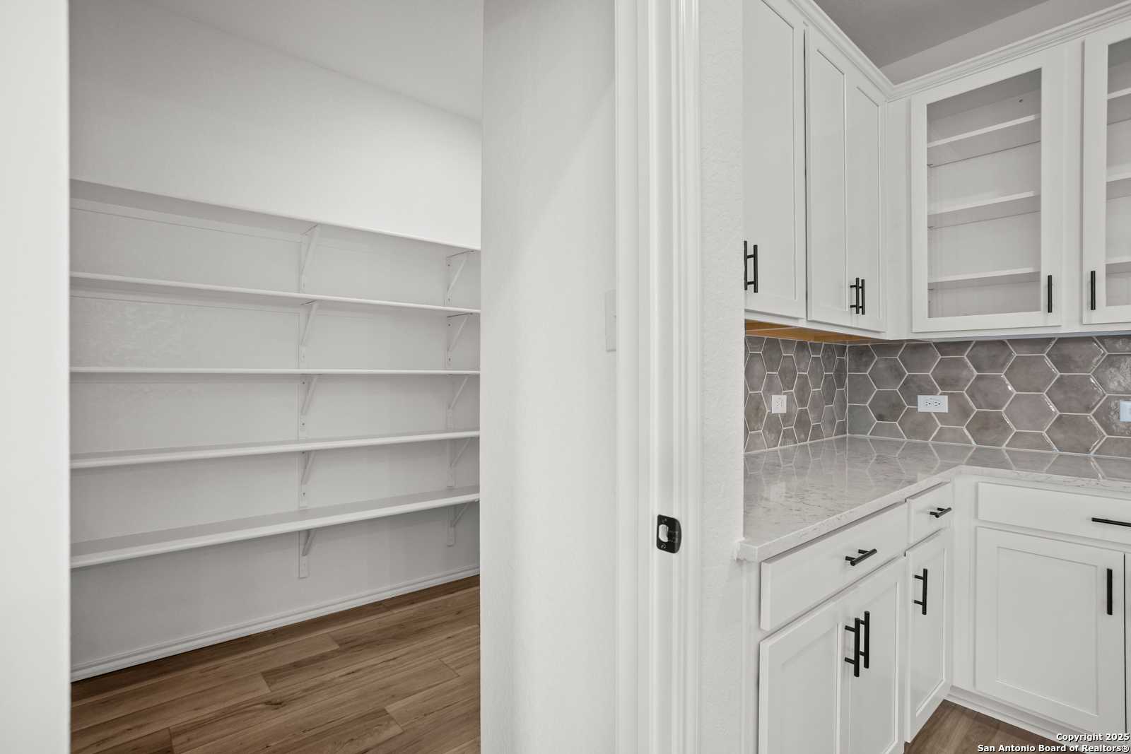 Modern white kitchen with quartz counters, gray hexagon backsplash, glass cabinets, and adjacent walk-in pantry in Davidson Homes The Daphne K, San Antonio