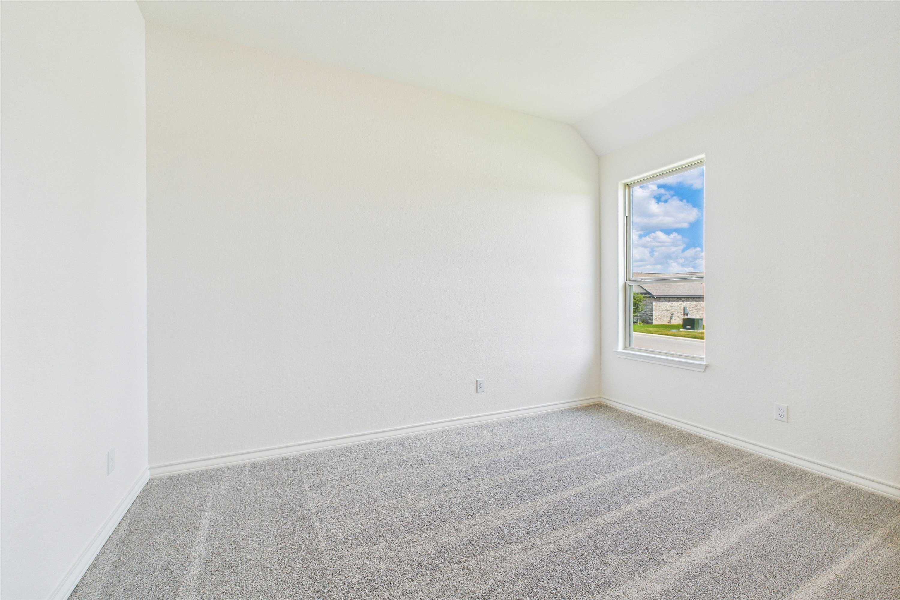 Bright empty bedroom with white walls, beige carpet, and large window showing blue sky in 4-bedroom Summerlin C home, Castroville, TX