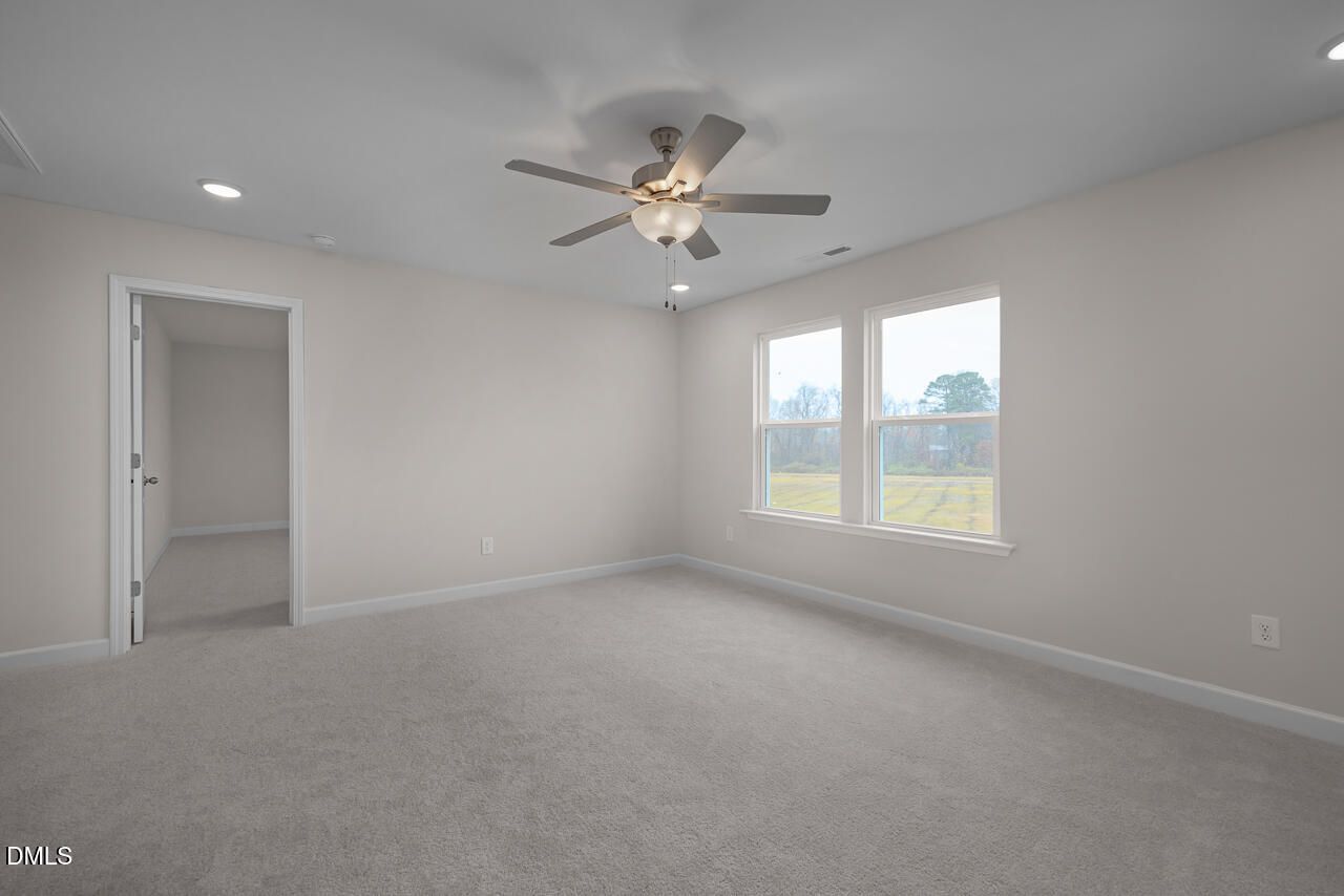 Bright empty bedroom with ceiling fan, beige walls, gray carpet, and large windows overlooking fields in The Cypress D II by Davidson Homes, Angier, NC