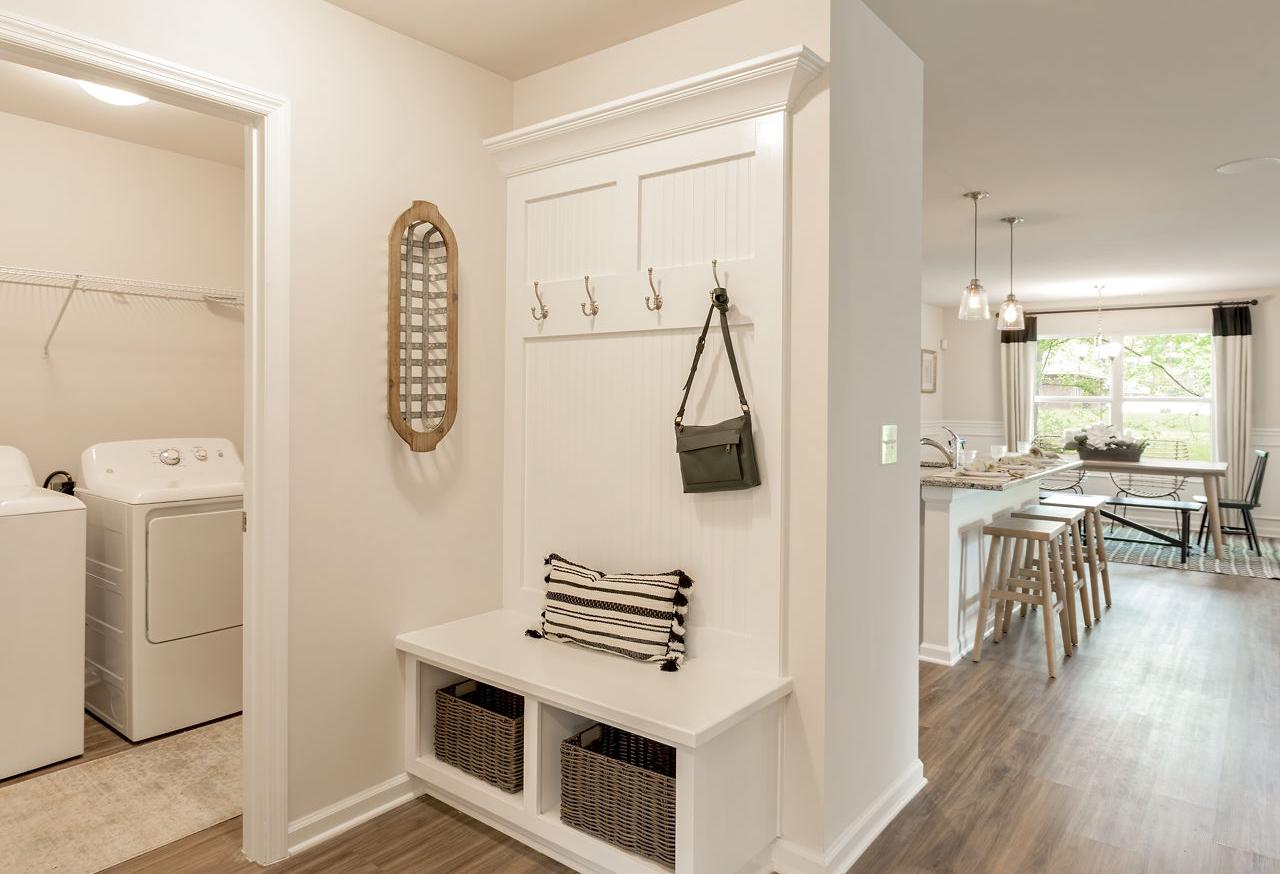 Spacious mudroom laundry in The Franklin home design with white washer dryer, built-in bench hooks, baskets, and open kitchen view