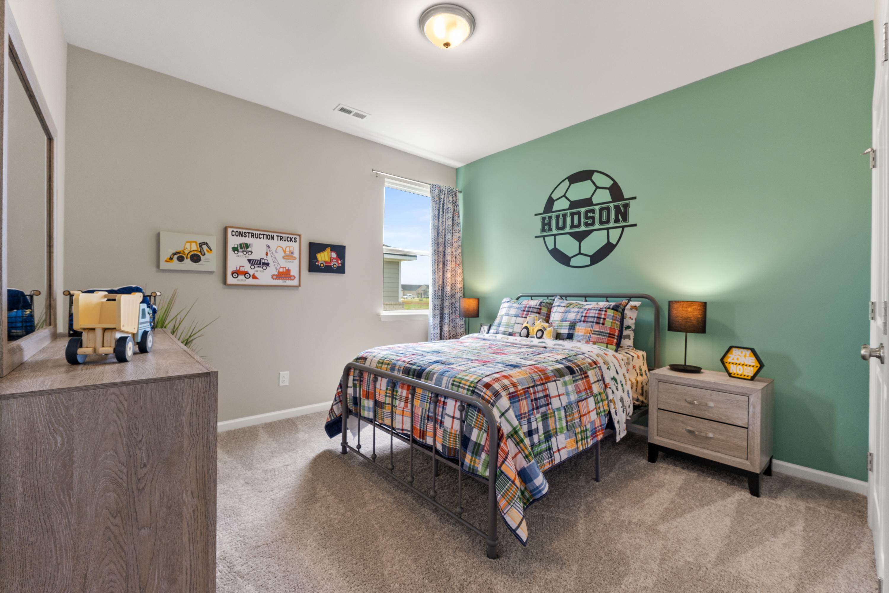 Boy's soccer-themed bedroom at The Meadows at Hampton Cove in Owens Cross Roads AL with green accent wall, colorful plaid bedding, and toy displays