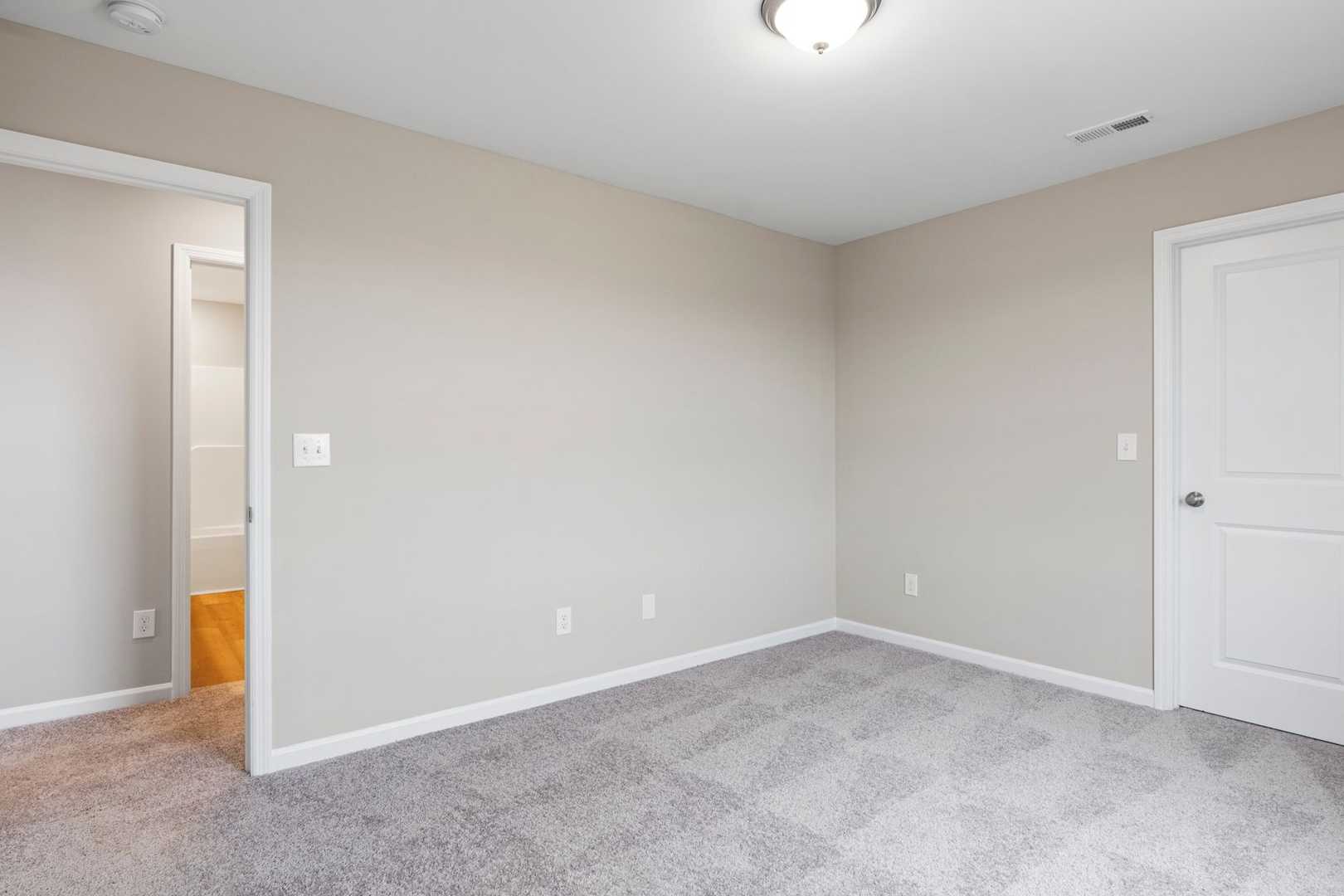Spacious bedroom in The Stella home design featuring beige walls, gray carpet flooring, and adjacent bathroom doorway
