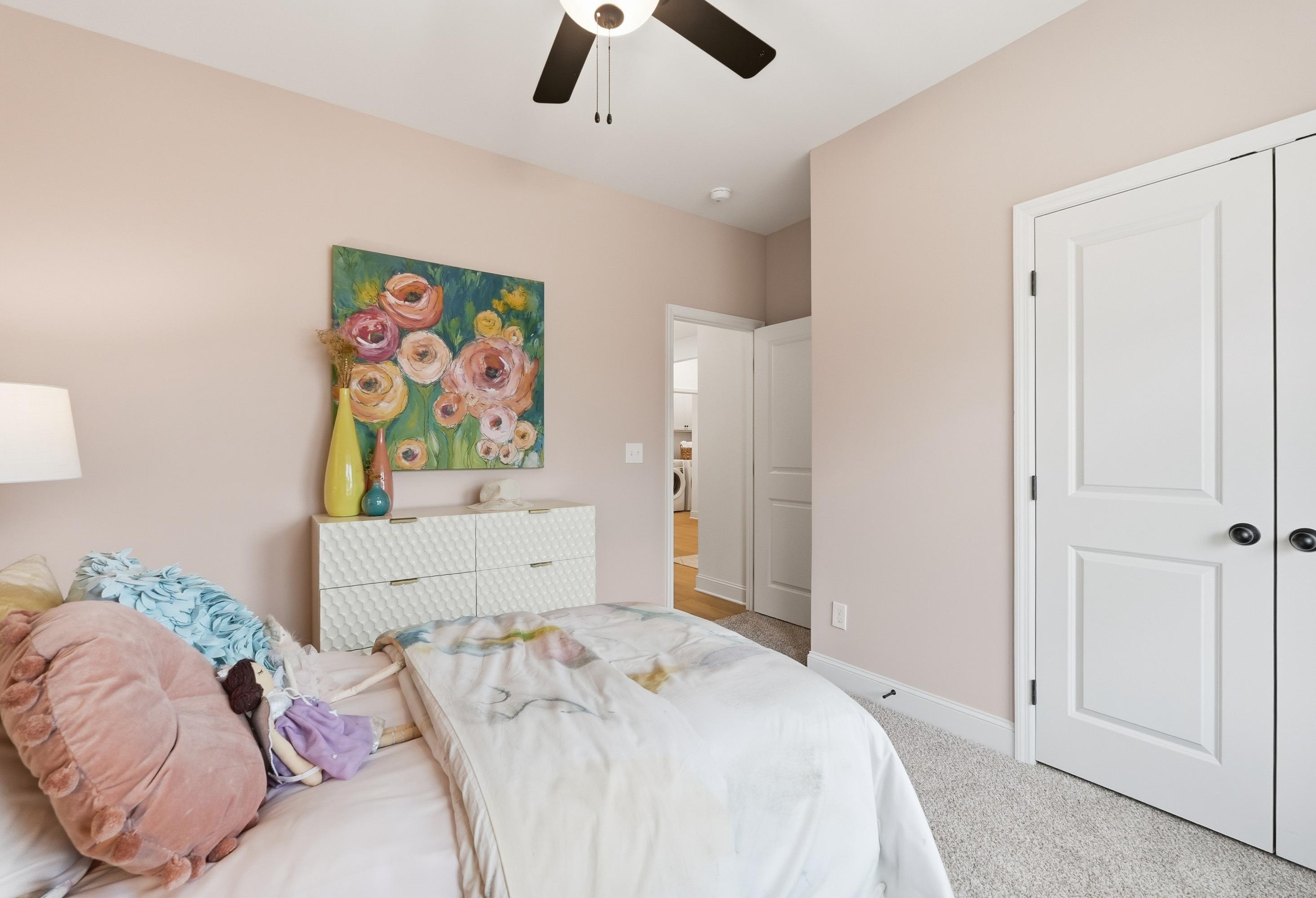 Cozy master bedroom in The Laurel with pink walls, floral artwork, ceiling fan, white doors, and plush bed