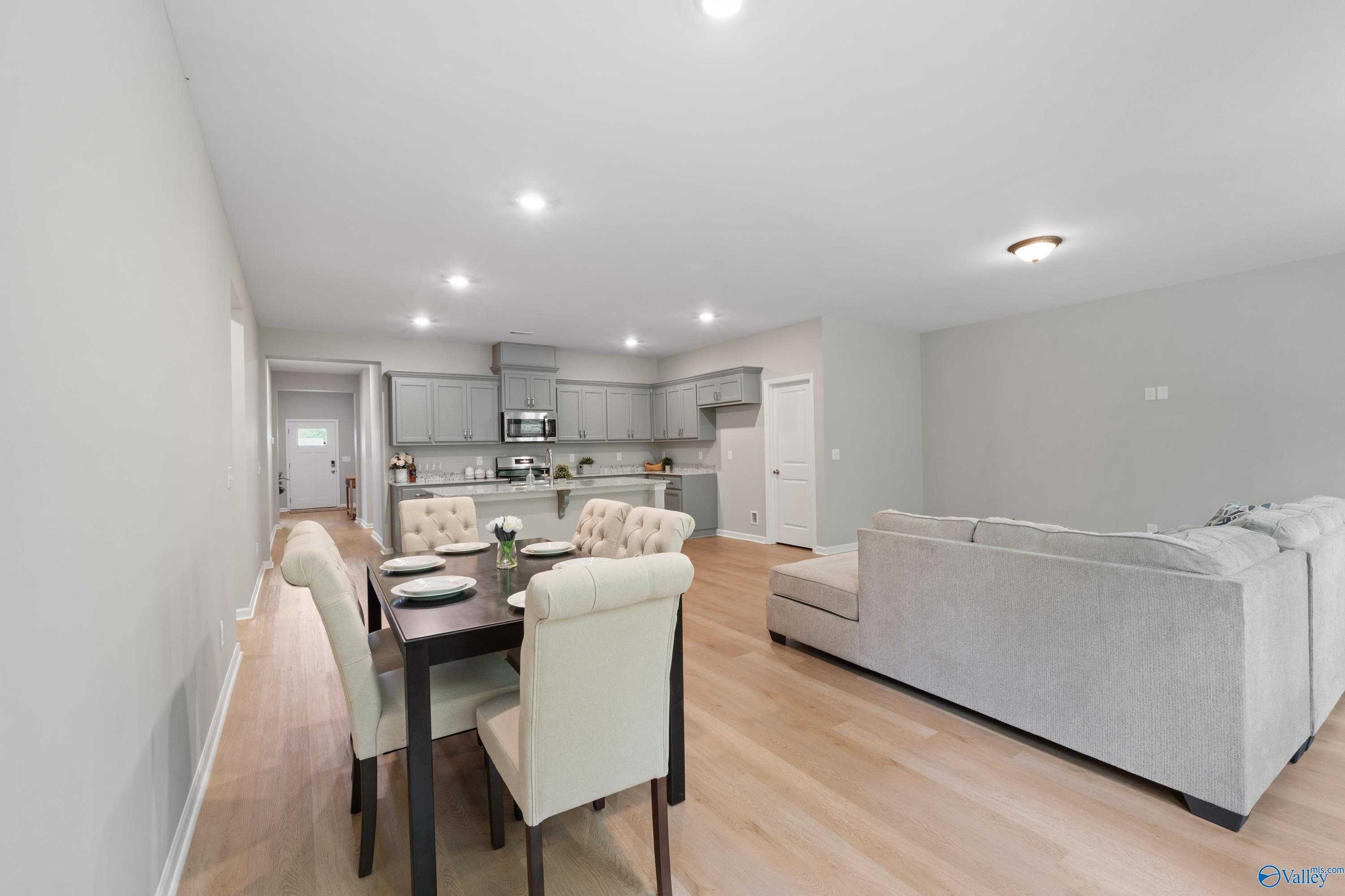 Open-concept living dining area with gray L-shaped sofa, wooden table, beige chairs, hardwood floors in Davidson Homes Luna, Hazel Green, AL