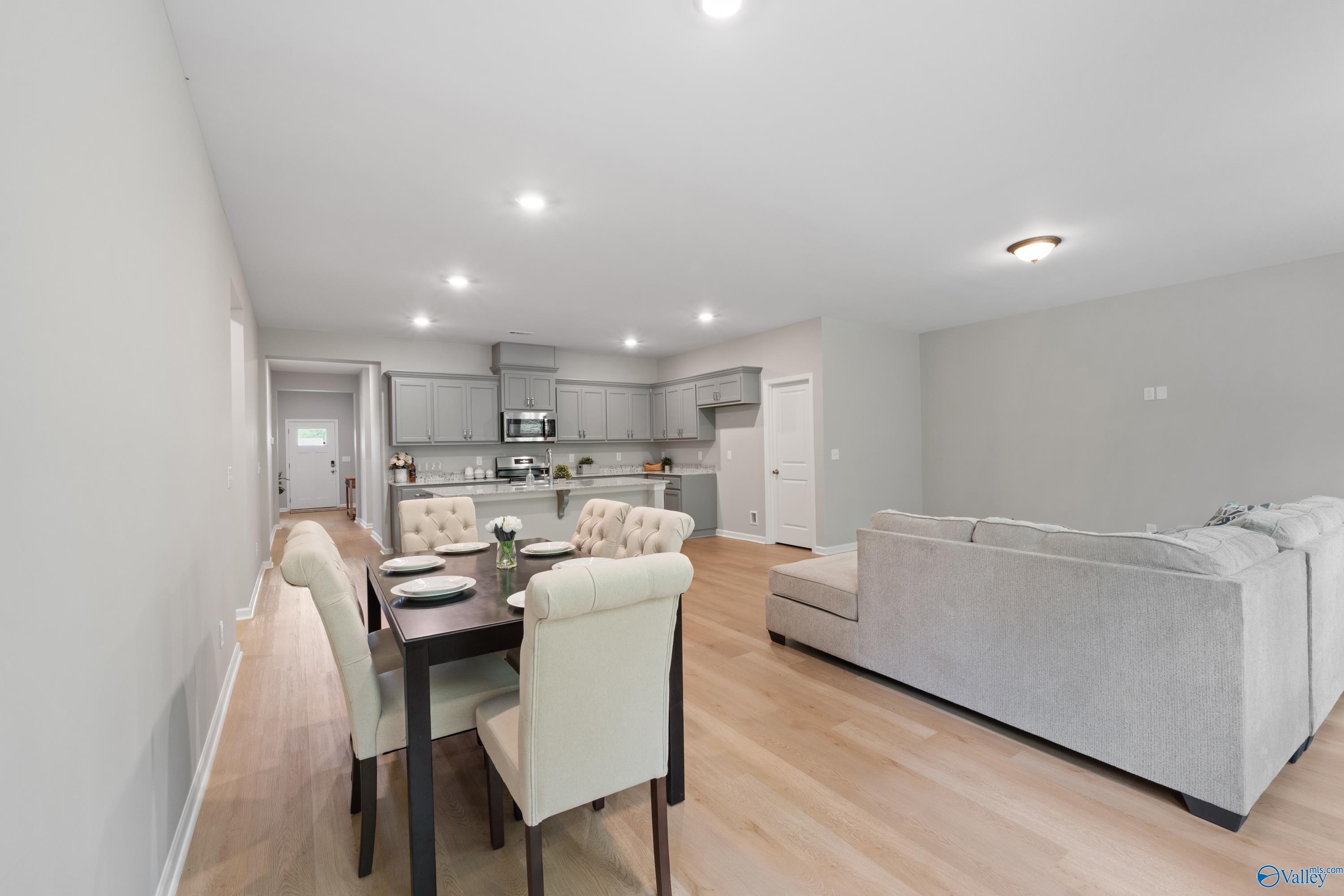 Spacious open-concept living-dining area with gray sectional sofa, wooden table, beige chairs, and adjacent kitchen in Davidson Homes The Luna, Hazel Green, AL