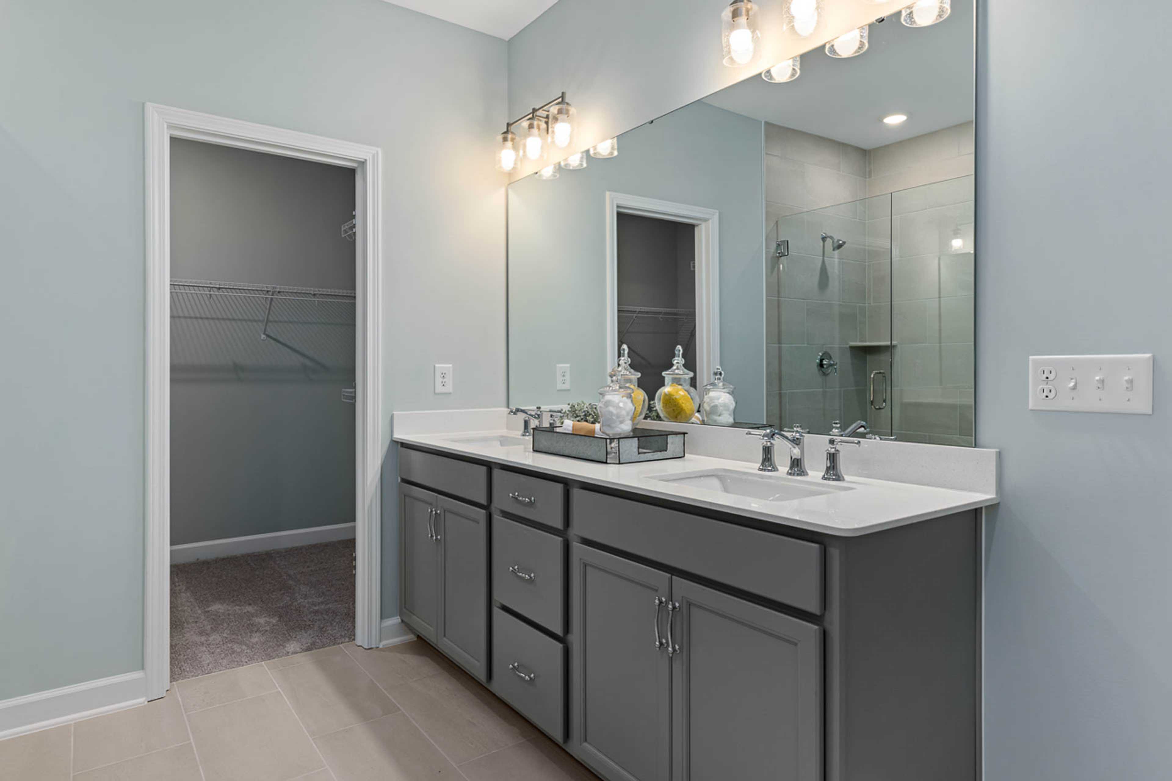 Spacious primary bathroom at Weatherford East in Angier NC with gray double vanity, glass shower, walk-in closet, and soft blue walls