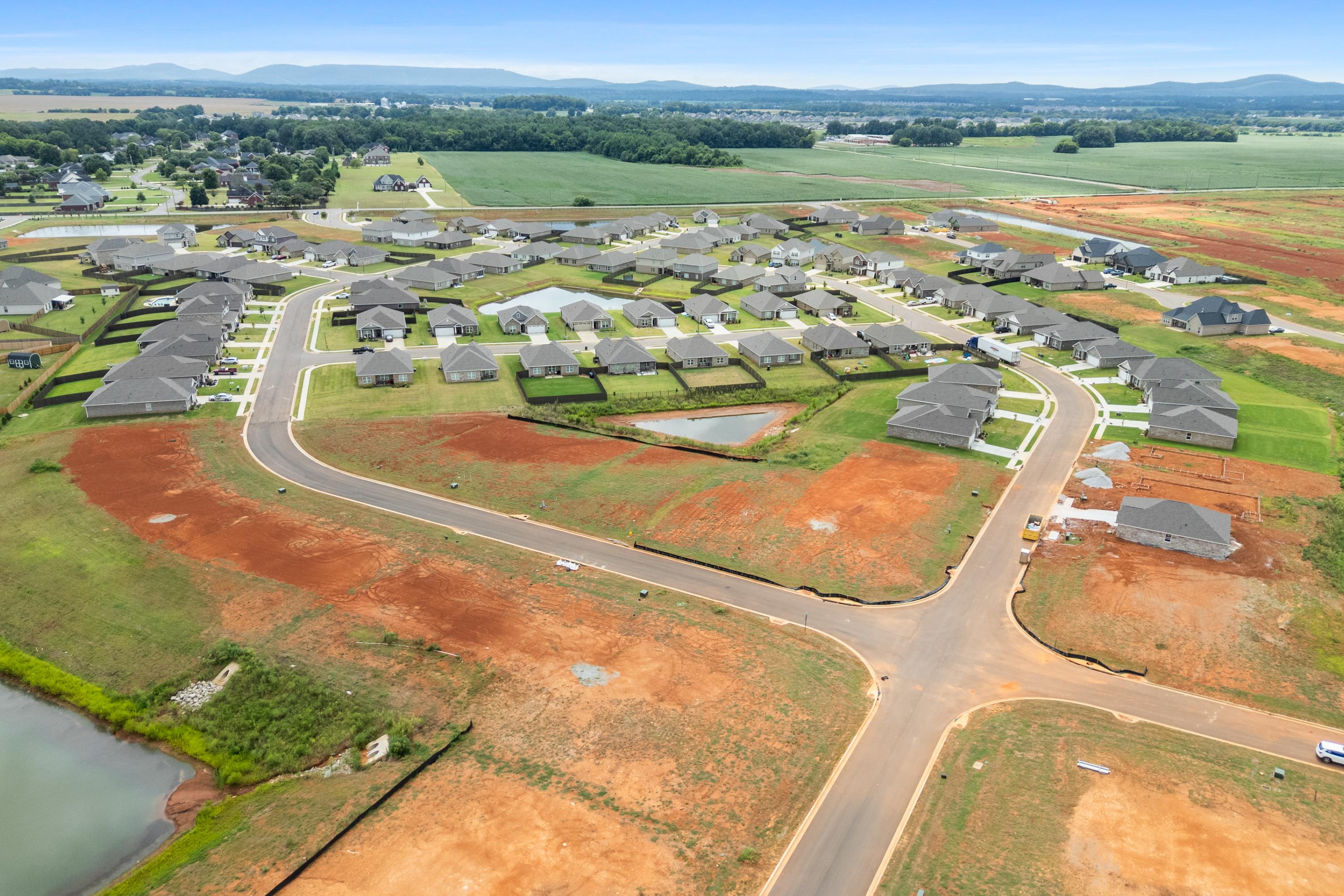 Aerial view of Heritage Lakes in New Market AL by Davidson Homes with new clustered homes, curved roads, red dirt lots, green fields, and pond