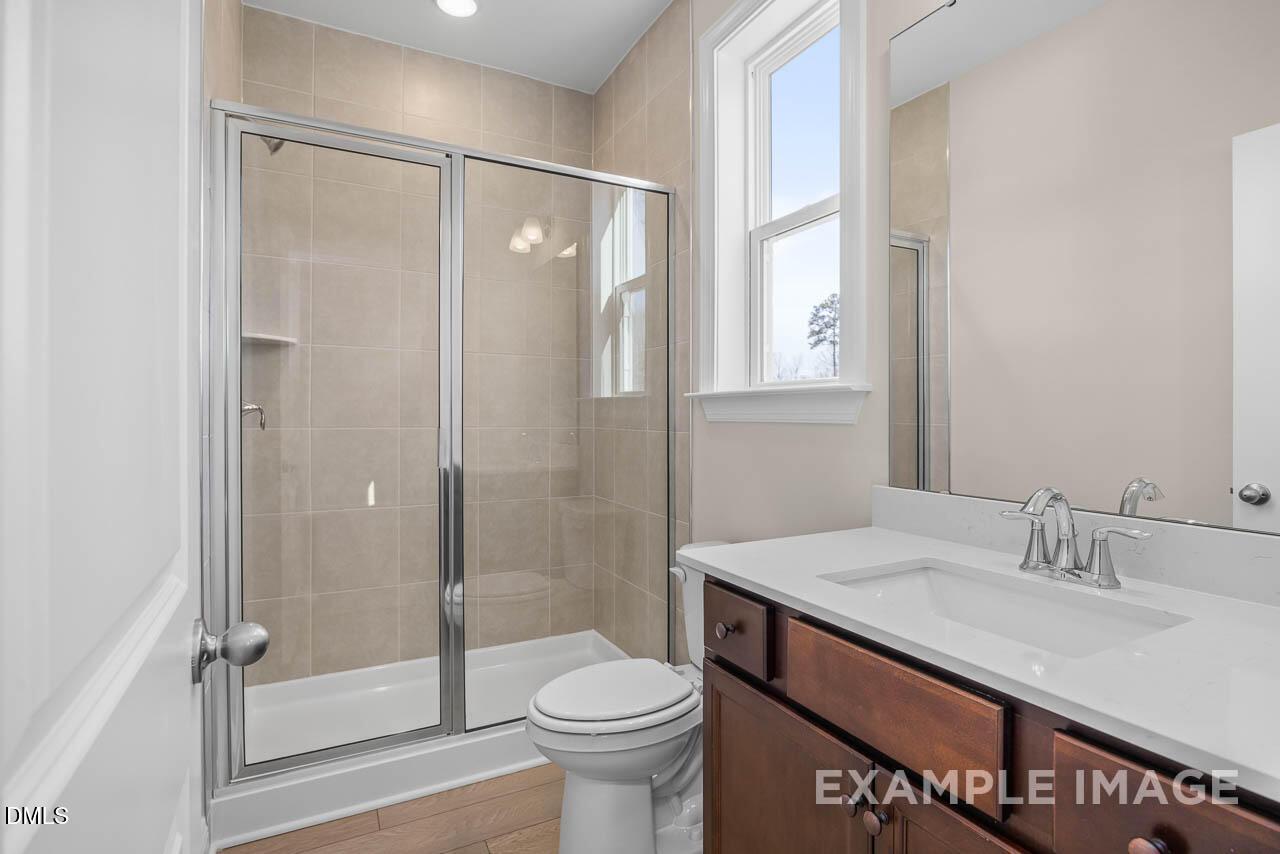 Elegant bathroom with frameless glass shower, beige tile walls, wood vanity, and natural light in Crawford D home, Angier, NC