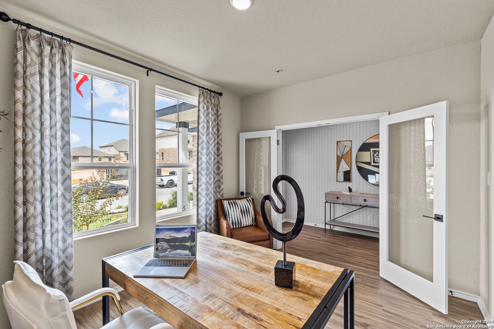Modern home office with wooden desk, laptop, abstract sculpture, and large windows overlooking neighborhood in Davidson Homes The Douglas F, Bricewood, San Antonio