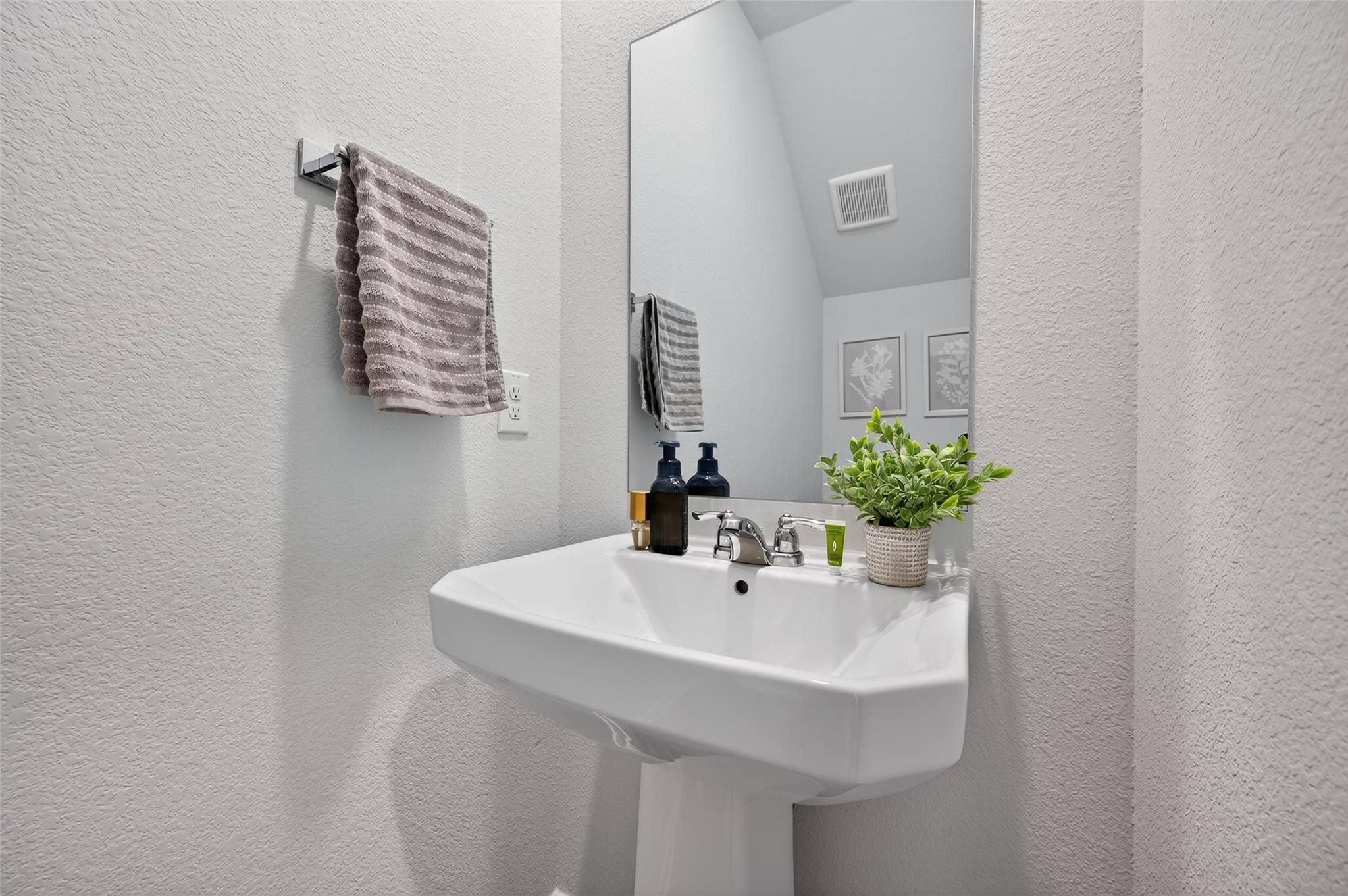 Modern guest bathroom with white pedestal sink, large mirror, hanging towels, and potted plant in Davidson Homes The Brazos E, Magnolia, Texas