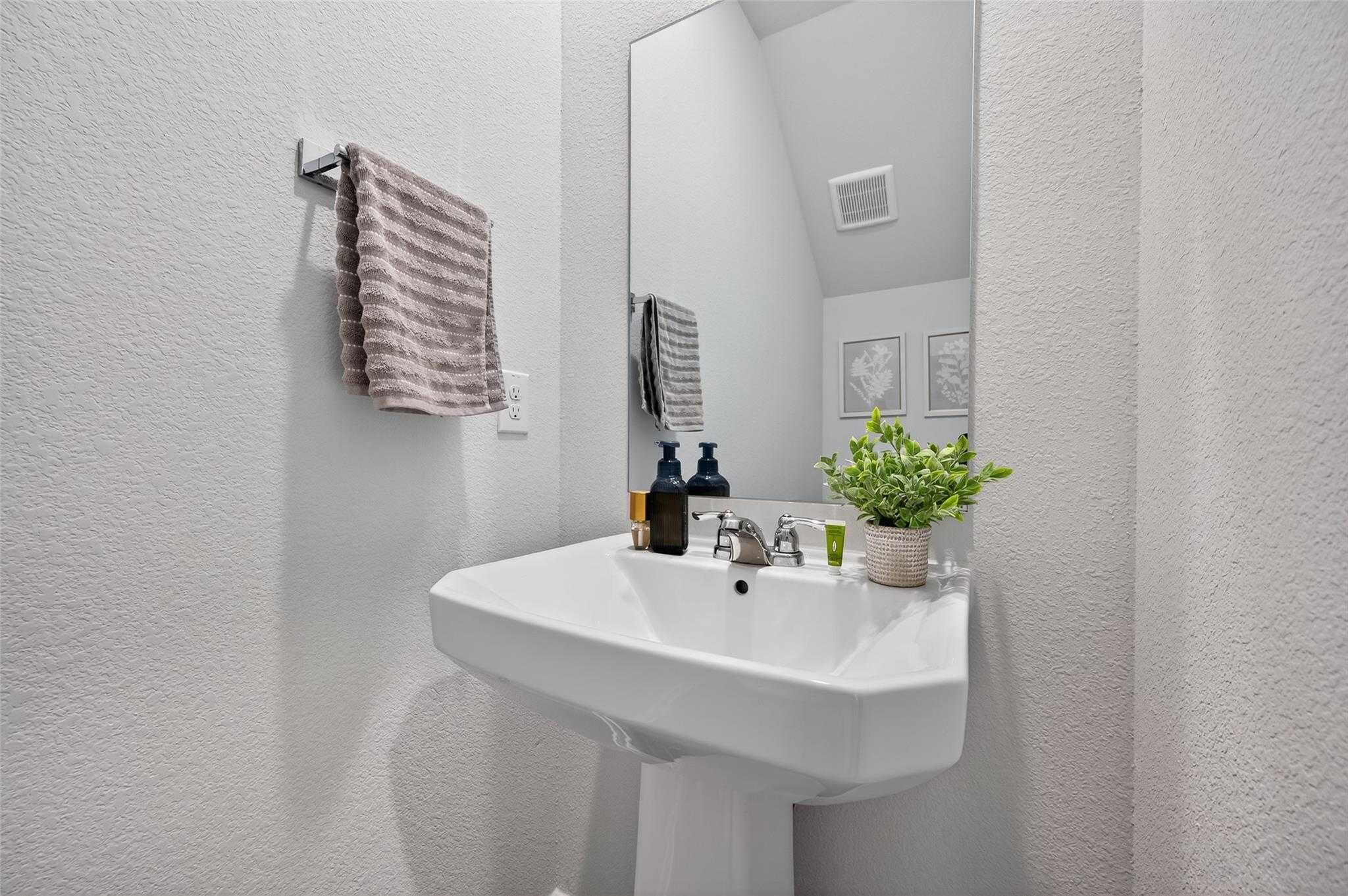 Modern guest bathroom with white pedestal sink, large mirror, hanging towels, and potted plant in Davidson Homes The Brazos E, Magnolia, Texas