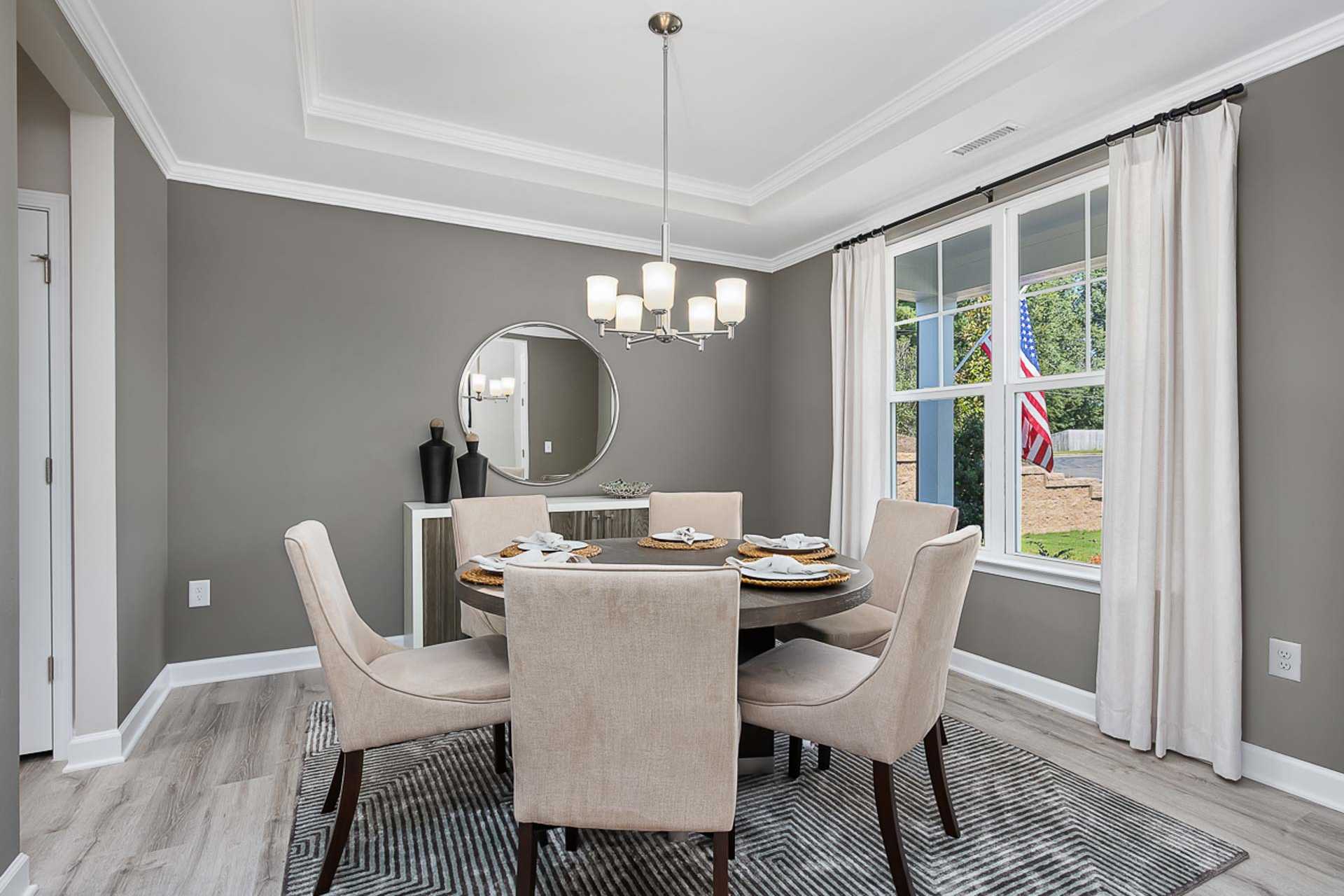 Elegant dining room at Prince Place in Fuquay-Varina NC with round wood table, beige upholstered chairs, modern chandelier, gray walls, and window view