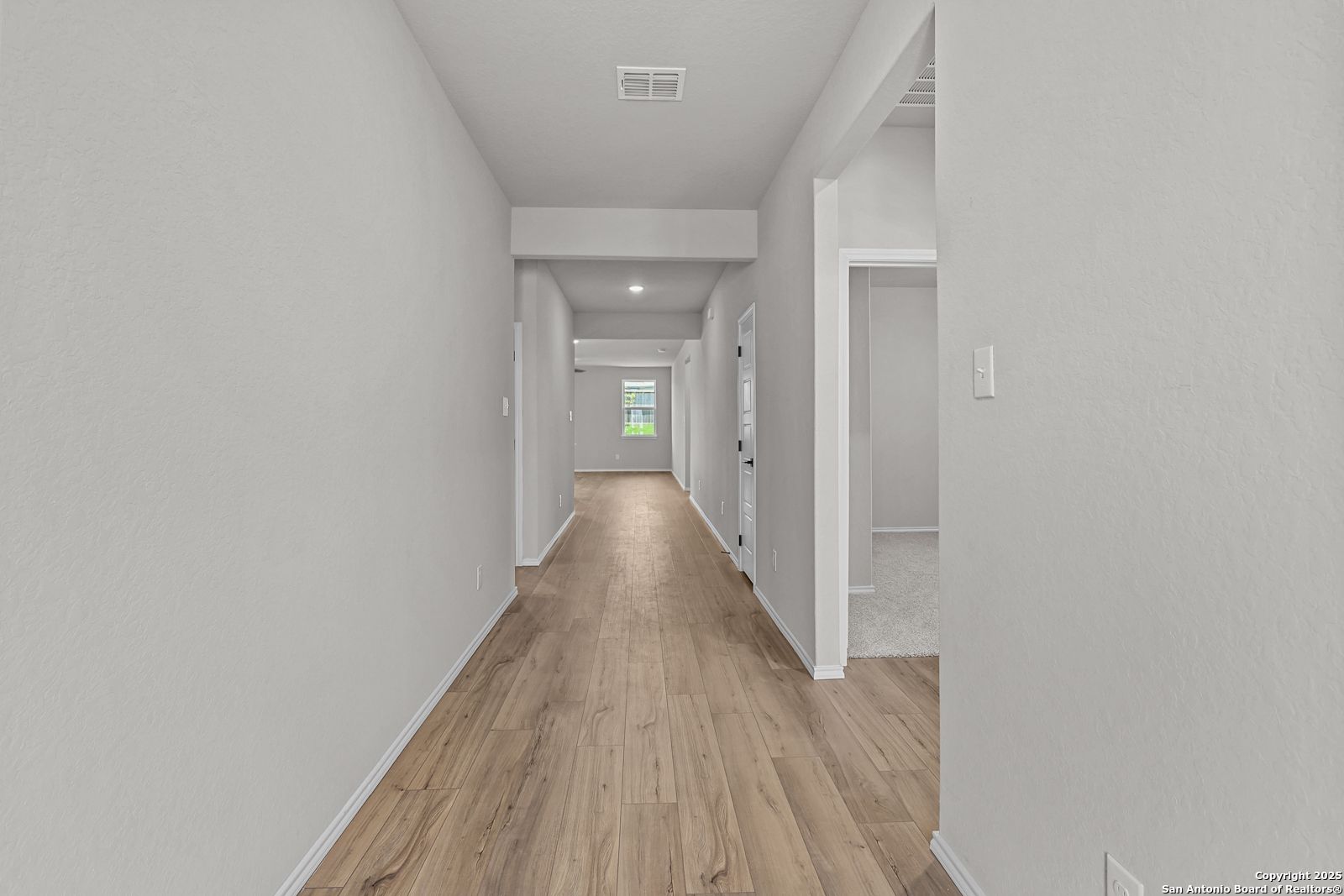 Long hallway with light wood floors and neutral walls leading to sunlit window in The Daphne K 4-bedroom home, San Antonio, Texas
