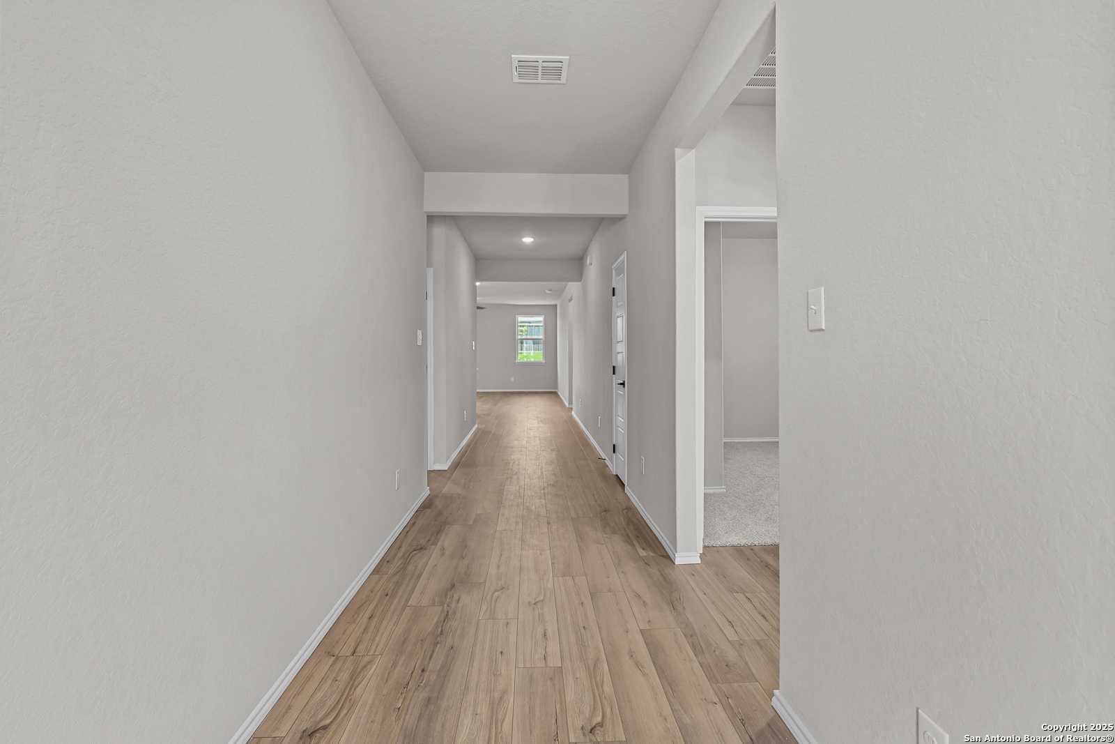 Long hallway with light wood floors and neutral walls leading to sunlit window in The Daphne K 4-bedroom home, San Antonio, Texas
