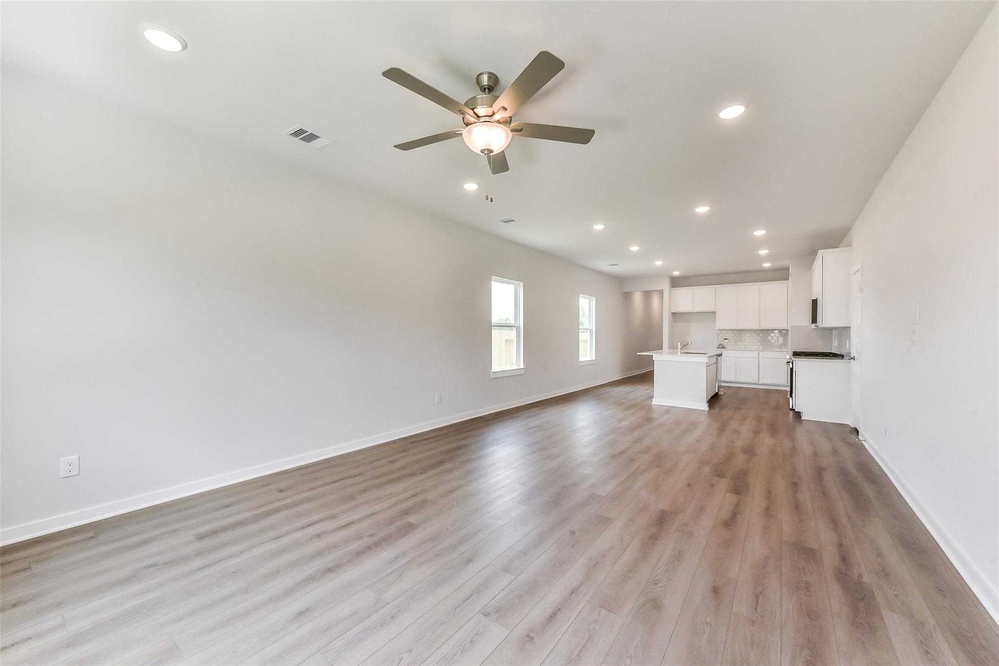 Spacious open living room with ceiling fan, wood laminate floors, and adjacent white cabinet kitchen in Davidson Homes The Frio F, Dayton, Texas
