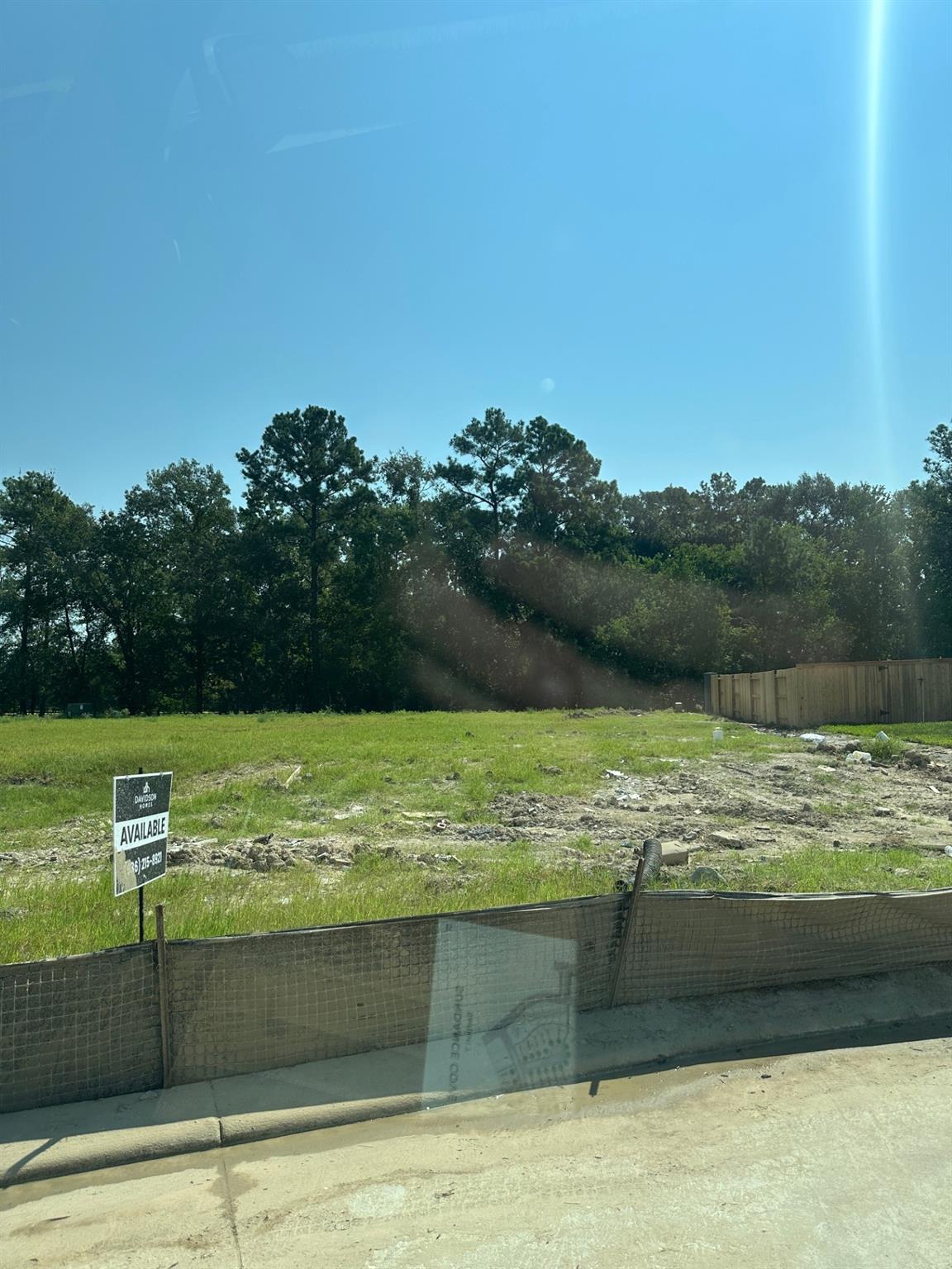 Vacant lot with Davidson Homes for-sale sign, silt fence, and pine trees under blue sky in Sundance Cove, Crosby, Texas