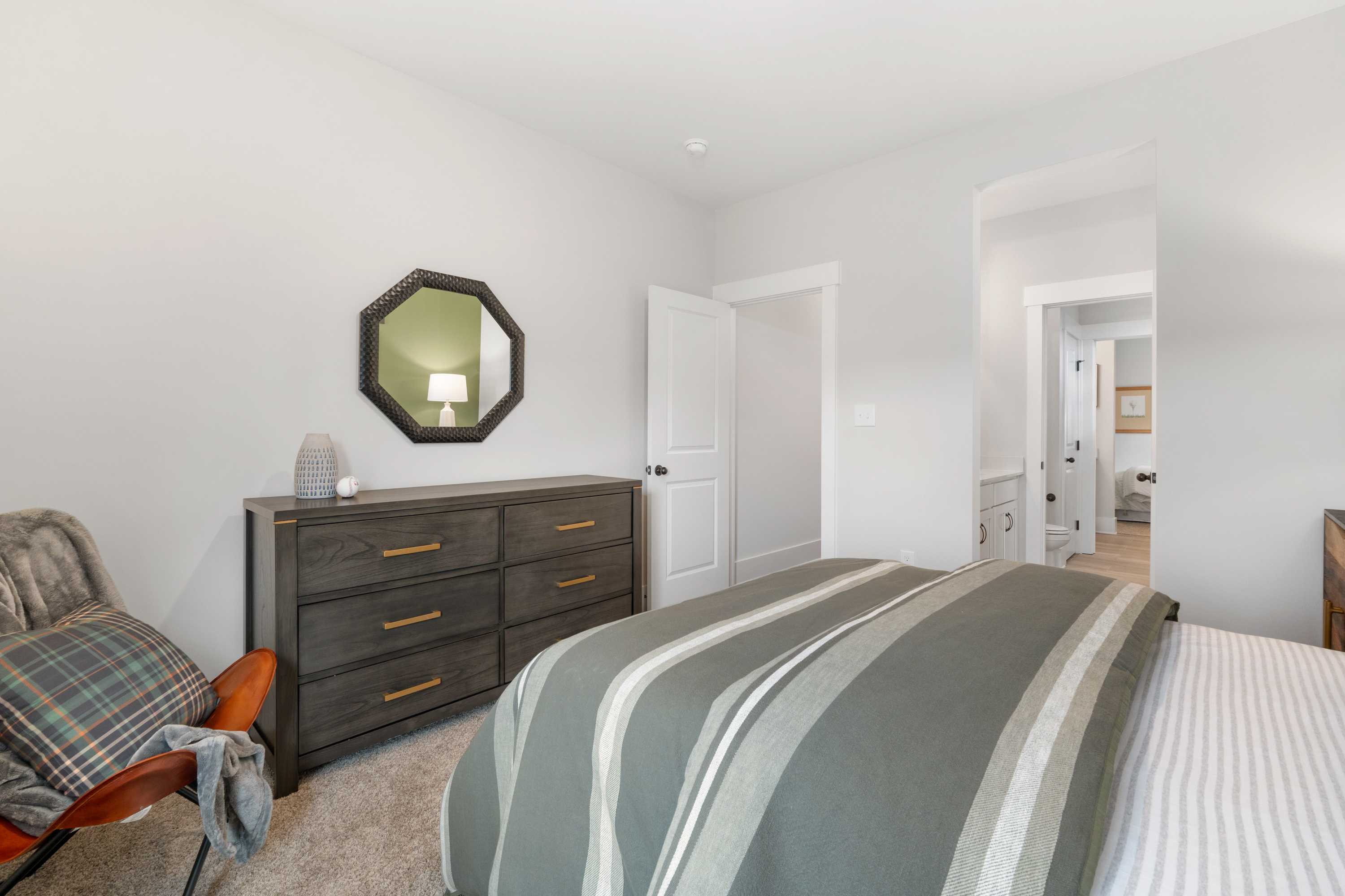 Cozy bedroom in Briercreek, Meridianville AL by Davidson Homes with queen bed, gray striped bedding, wooden dresser, orange chair, and round mirror