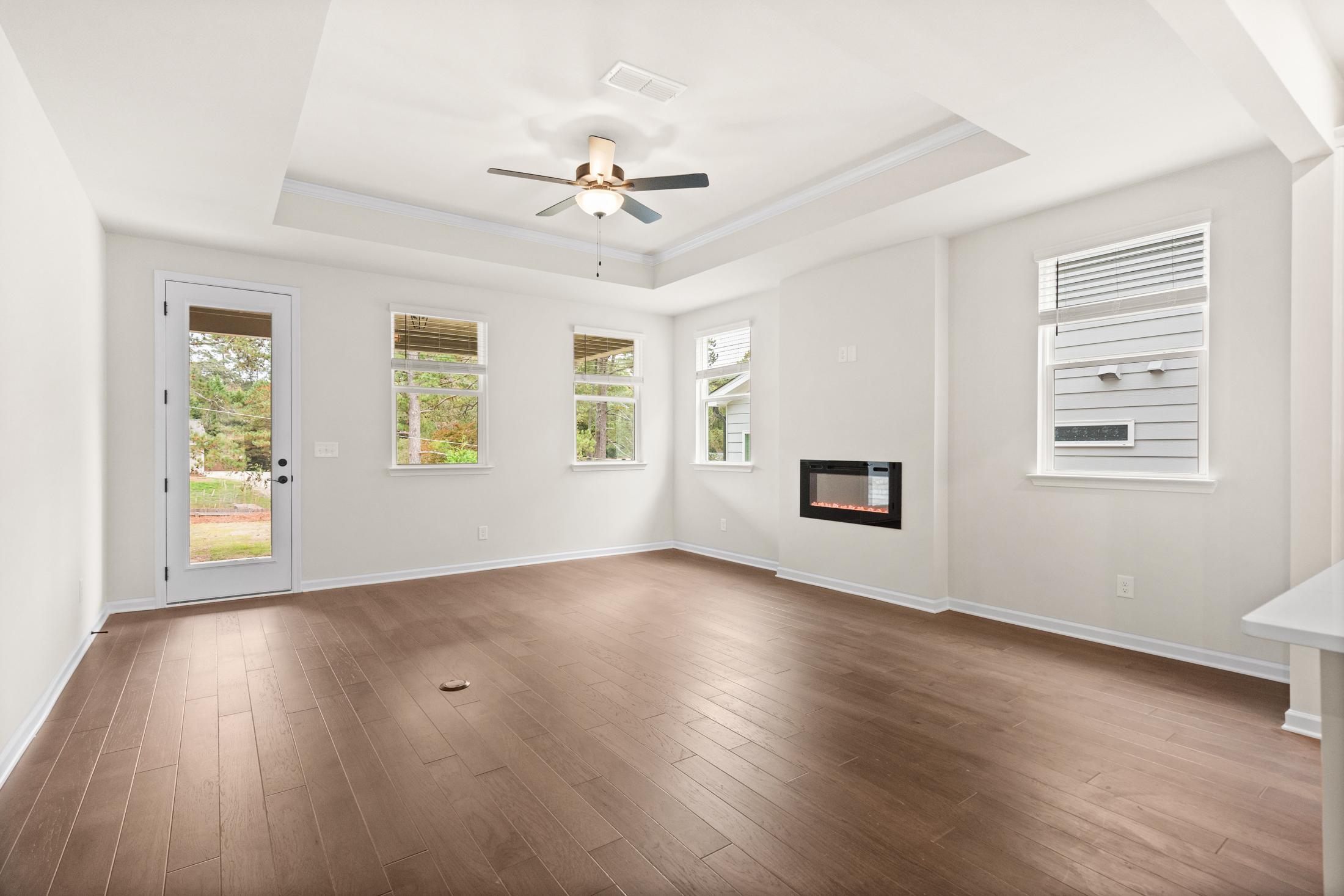 Spacious great room in The Glenwood home featuring gas fireplace, tray ceiling fan, hardwood floors, and large windows