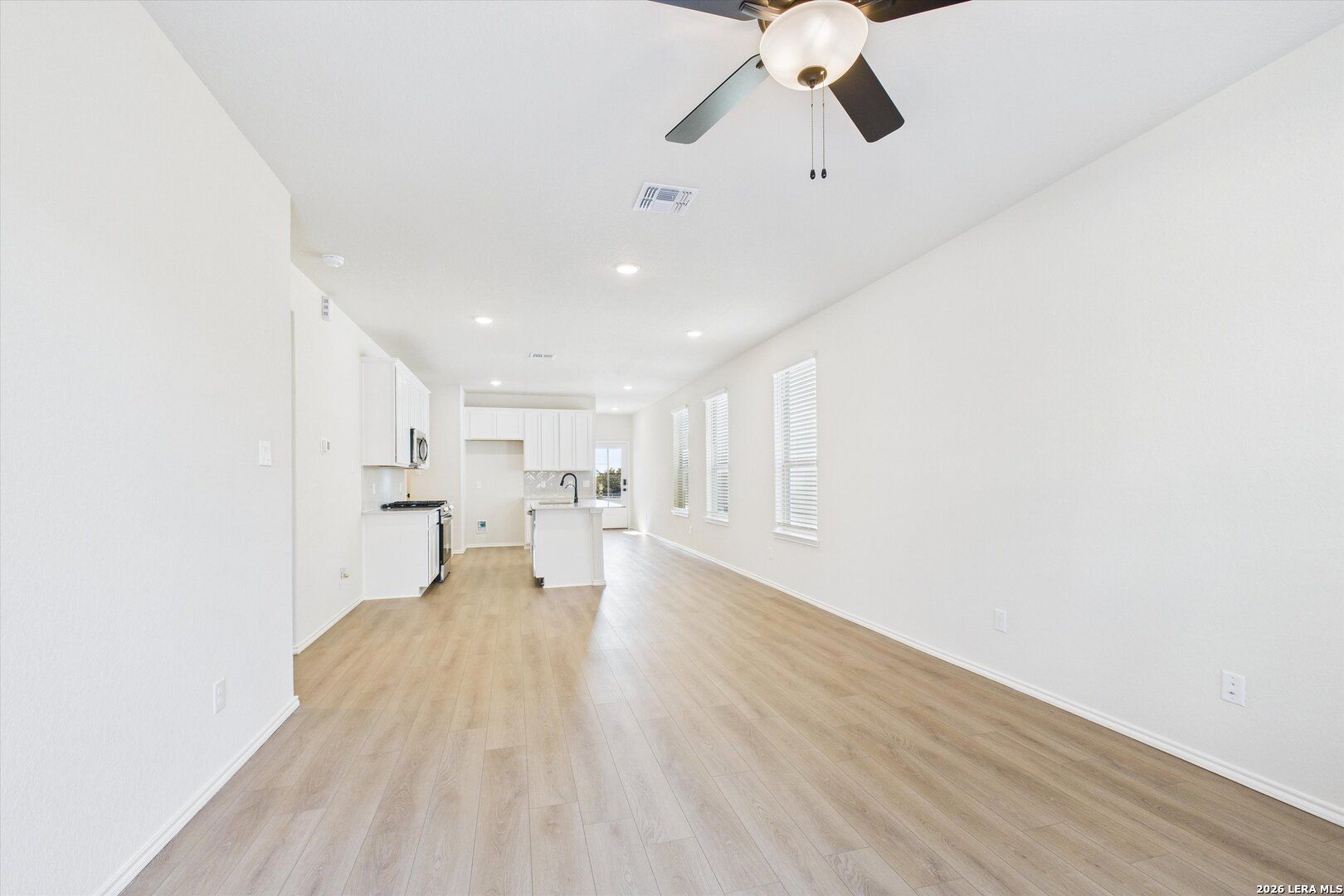 Open-concept kitchen and living space with white cabinets, island, ceiling fan, and light wood flooring in The Gillian B, San Antonio home
