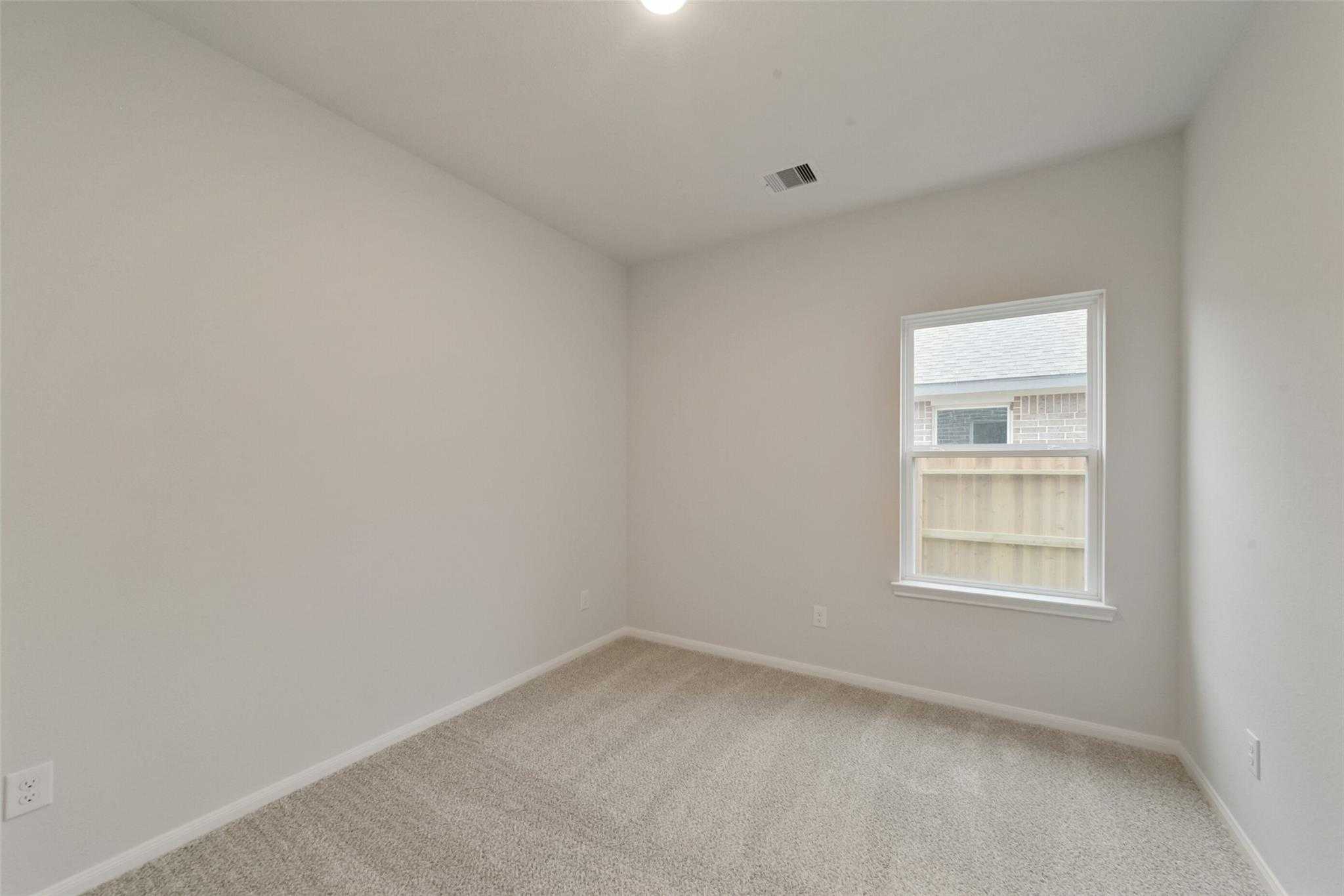 Empty secondary bedroom with neutral beige walls, carpet flooring, and window overlooking fence in Davidson Homes The San Marcos E, Beasley, Texas
