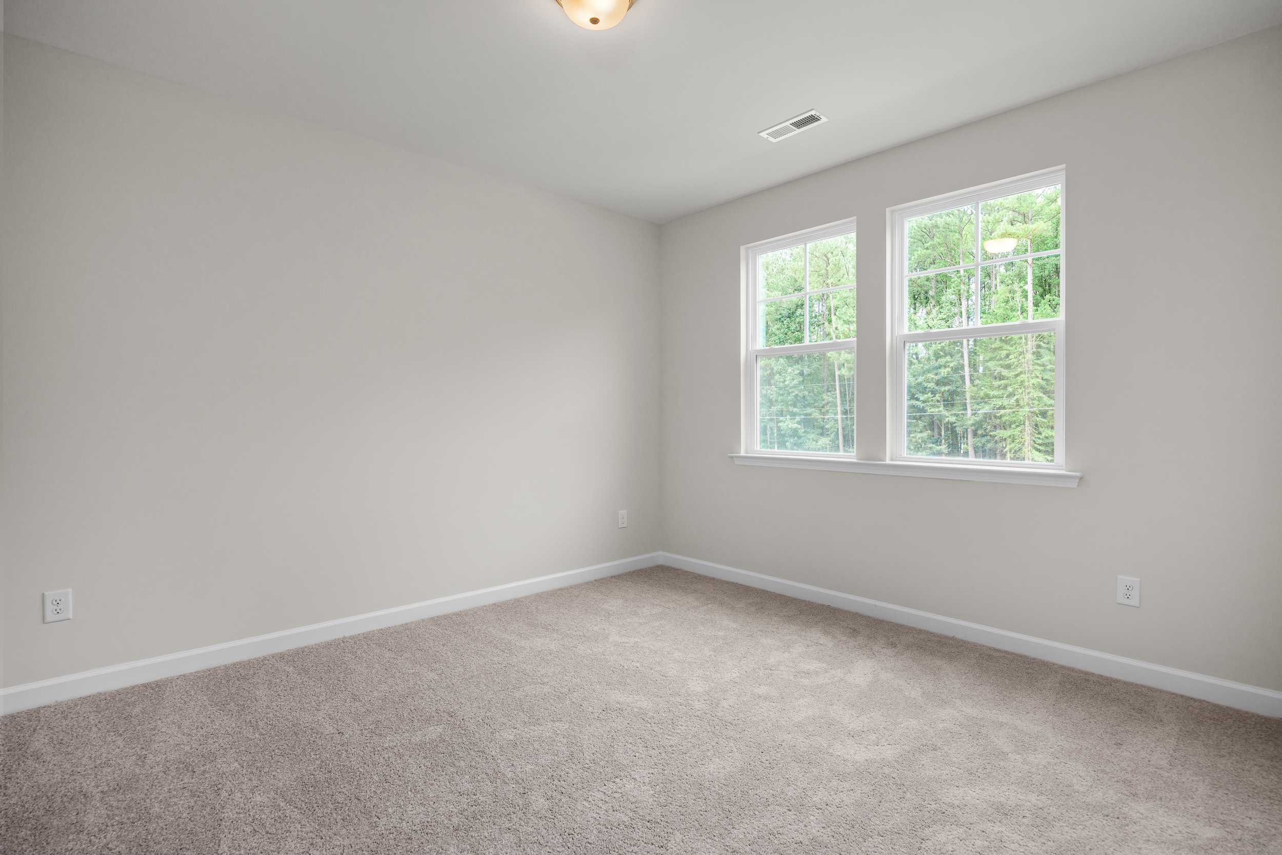 Spacious empty bedroom in The Aspen by Davidson Homes with neutral walls, large double windows, carpet flooring, and soft lighting