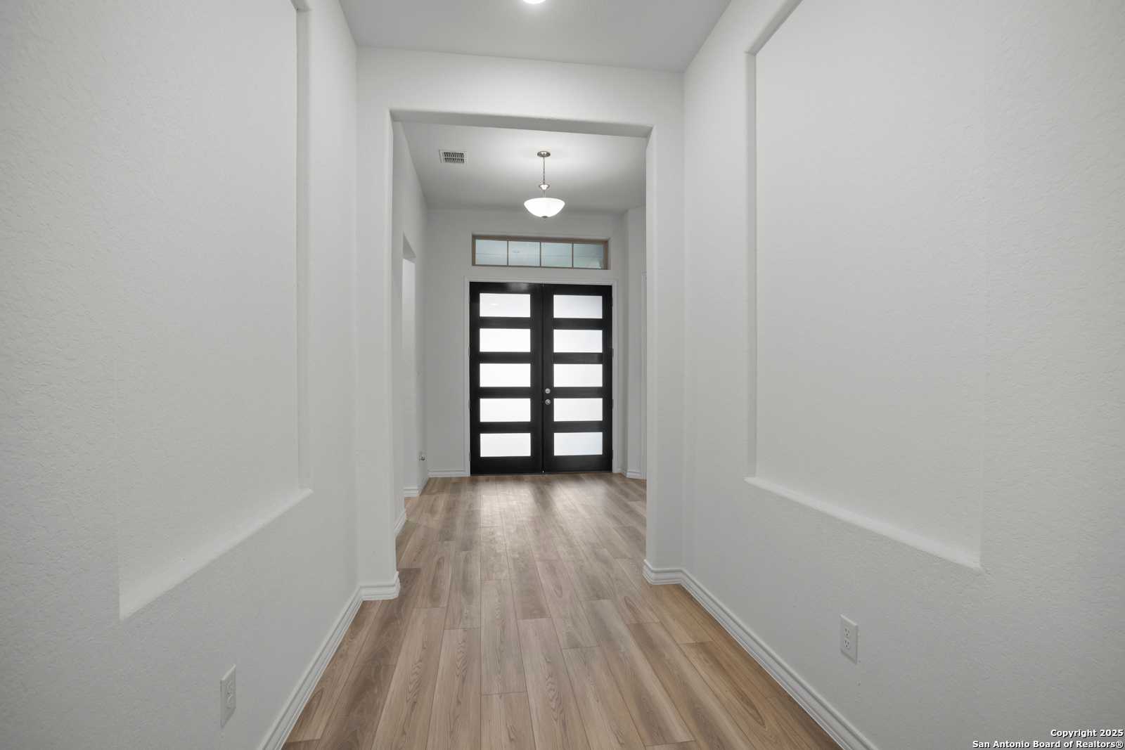 Elegant hallway with light oak floors, white walls, chandelier, and black glass French doors in Davidson Homes The Summerlin B, Castroville, Texas