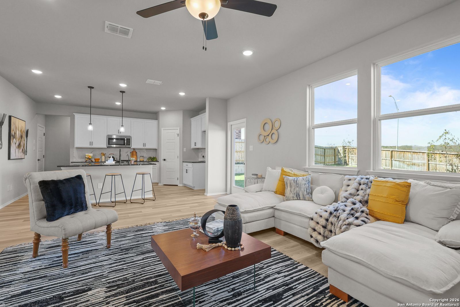 Bright open-concept living room with white L-shaped sofa, ceiling fan, and modern kitchen in Davidson Homes The Murray K, San Antonio