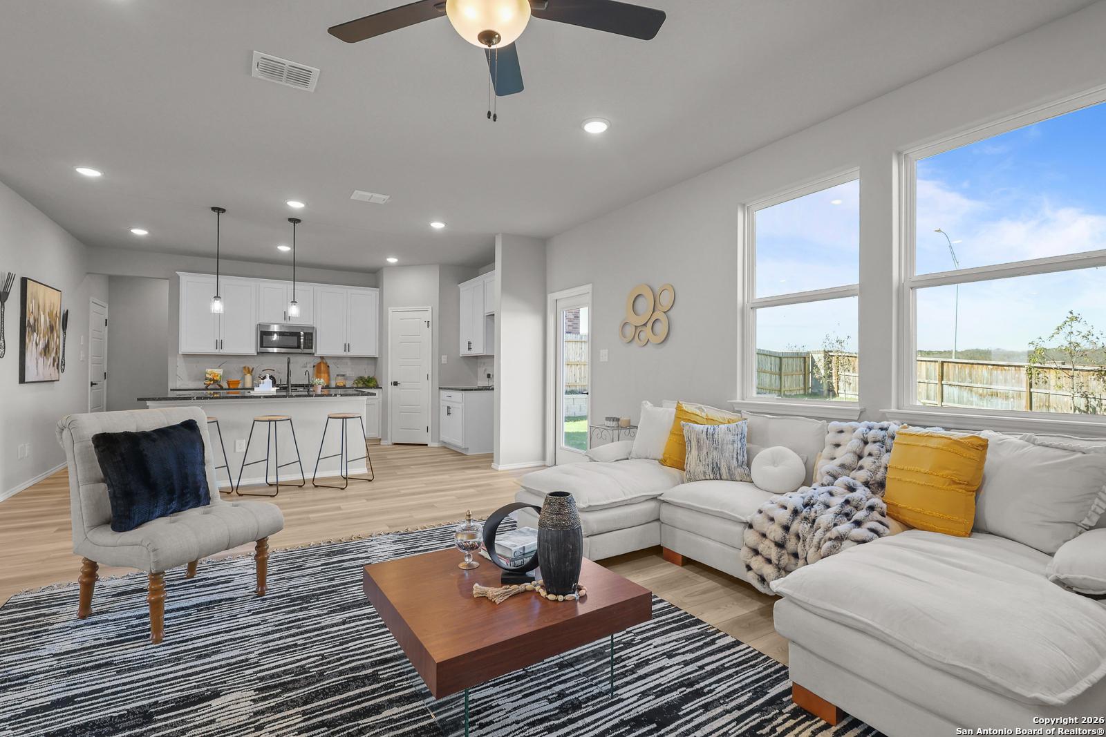 Bright open-concept living room with white L-shaped sofa, ceiling fan, and modern kitchen in Davidson Homes The Murray K, San Antonio