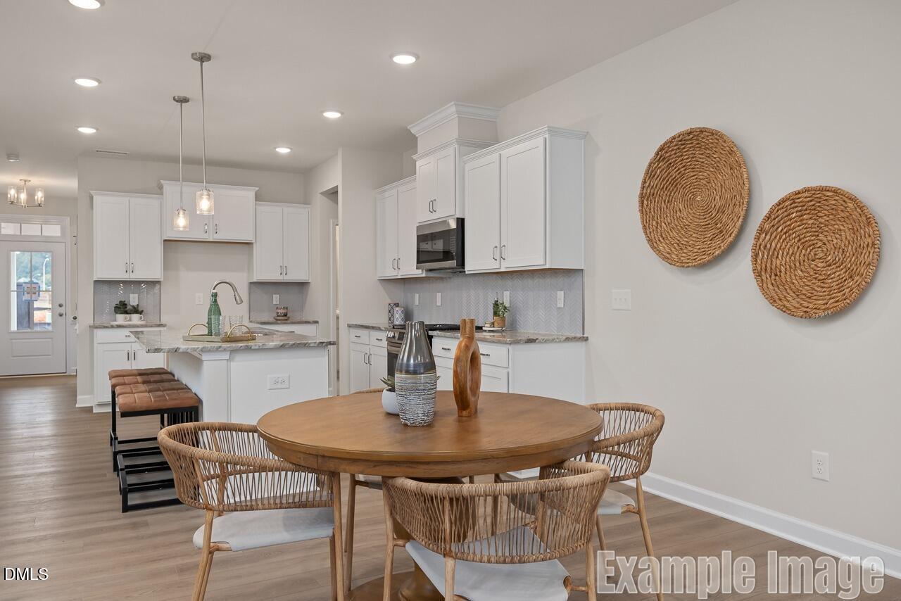 Bright open-concept kitchen with white cabinets, granite counters, stainless appliances, round wood dining table, and rattan chairs in The Carter C by Davidson Homes, Lillington, NC