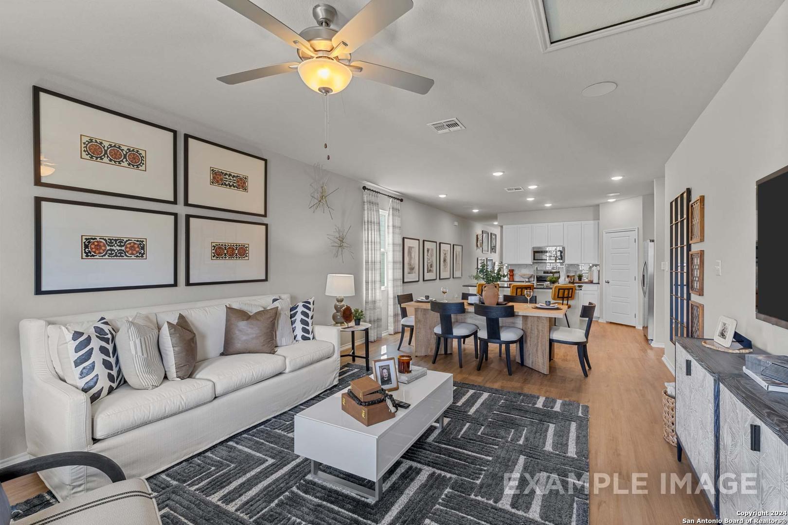 Open-concept living room with white sofa, dining table, ceiling fan, and adjacent white kitchen in The Sabine B by Davidson Homes, San Antonio