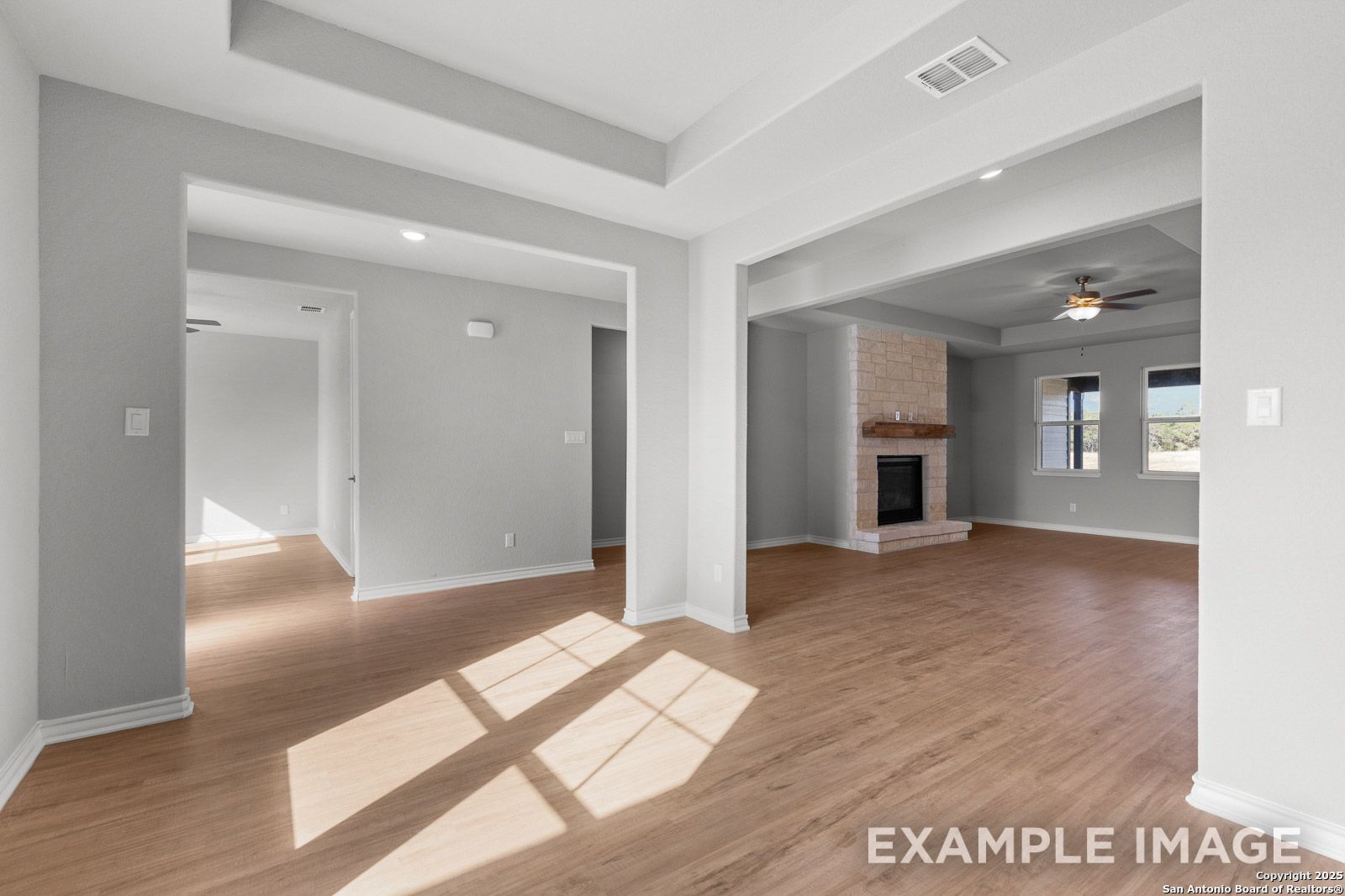 Open-concept living area in The Oxford featuring wood-look flooring, tray ceilings, and a stone fireplace in Castroville.