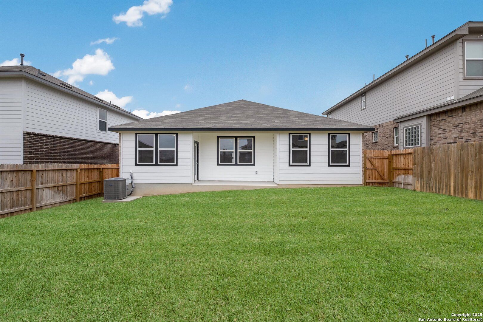 Spacious grassy backyard with wooden fence and rear view of white 1-story Asheville K home in Bricewood, San Antonio, Texas