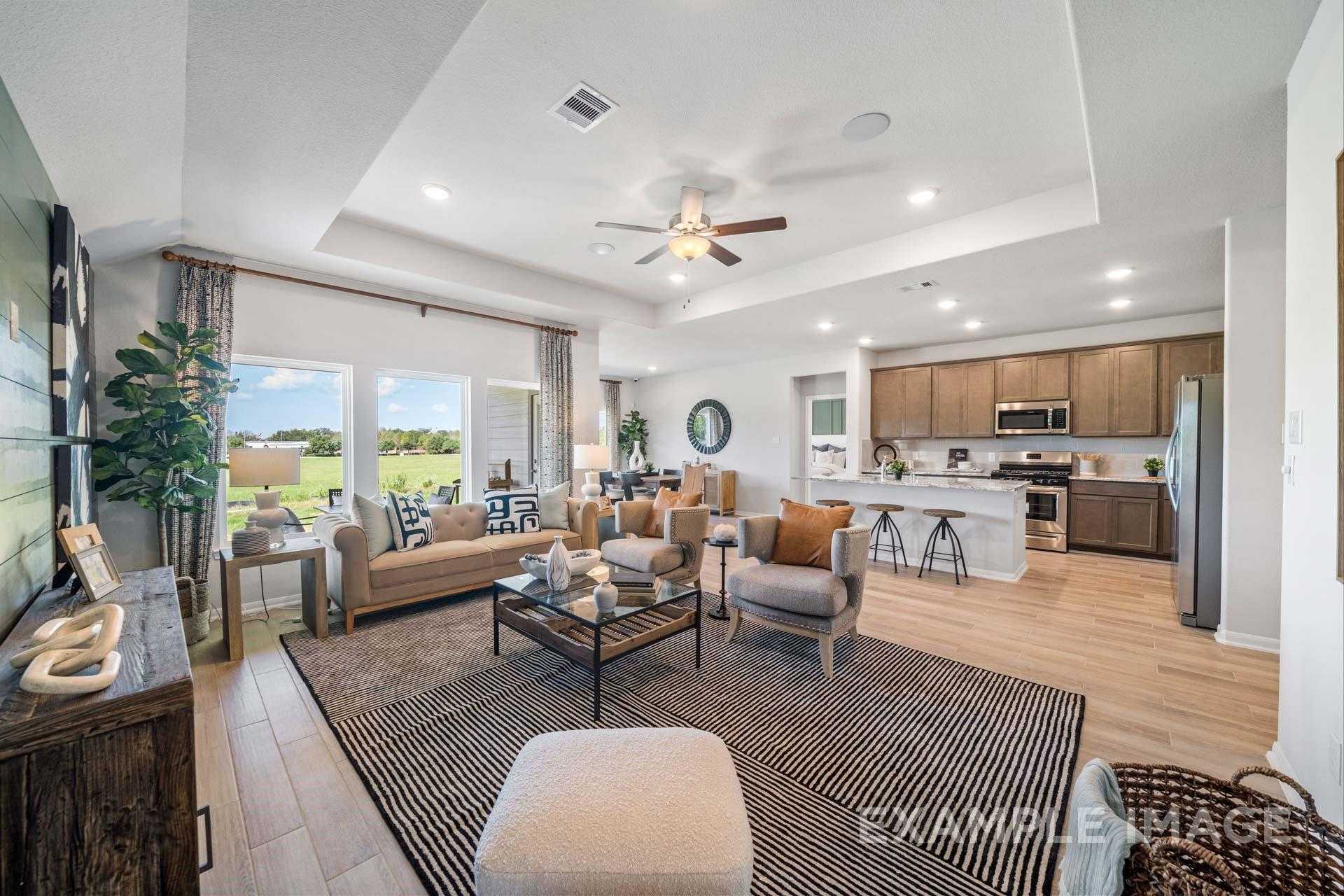 Open-concept living room and kitchen with beige sofa, wooden floors, and large windows overlooking green field in Davidson Homes The Acadia A, Lago Mar, Texas City