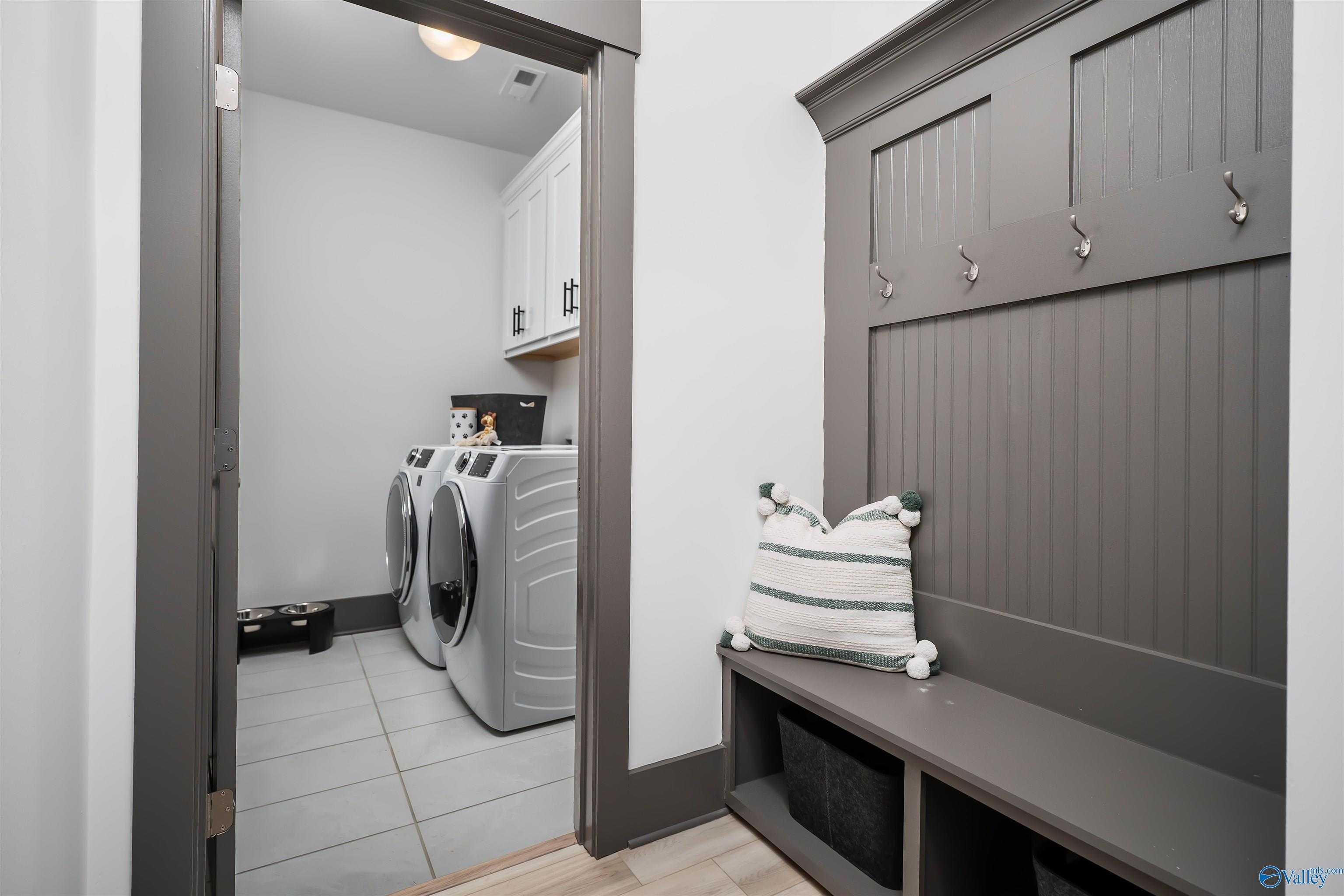 Modern laundry room featuring washer dryer, built-in gray bench with striped pillow, coat hooks in Davidson Homes The Rockford B, Madison AL