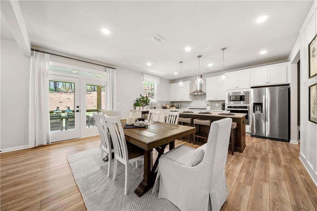 Open-concept dining room with rustic wooden table, white chairs, and adjacent kitchen with white cabinets, stainless appliances in Davidson Homes The Willow B, Dallas, GA