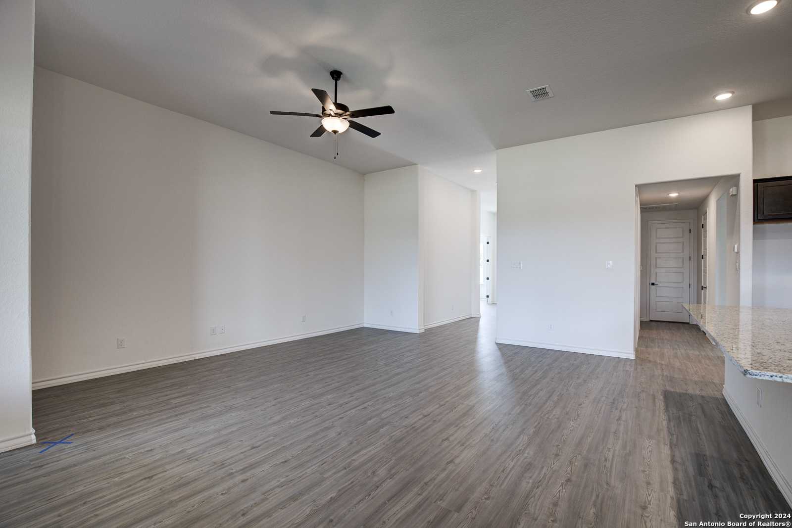 Open living room with ceiling fan, wood floors, and white walls flowing to modern kitchen in Davidson Homes The Garner B, Castroville, Texas