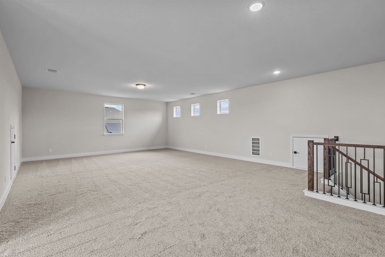 Spacious carpeted game room in The Belmont home design featuring a wrought iron staircase and ample natural light.