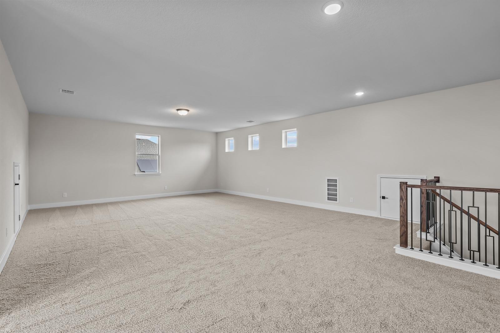 Spacious carpeted game room in The Belmont home design featuring a wrought iron staircase and ample natural light.